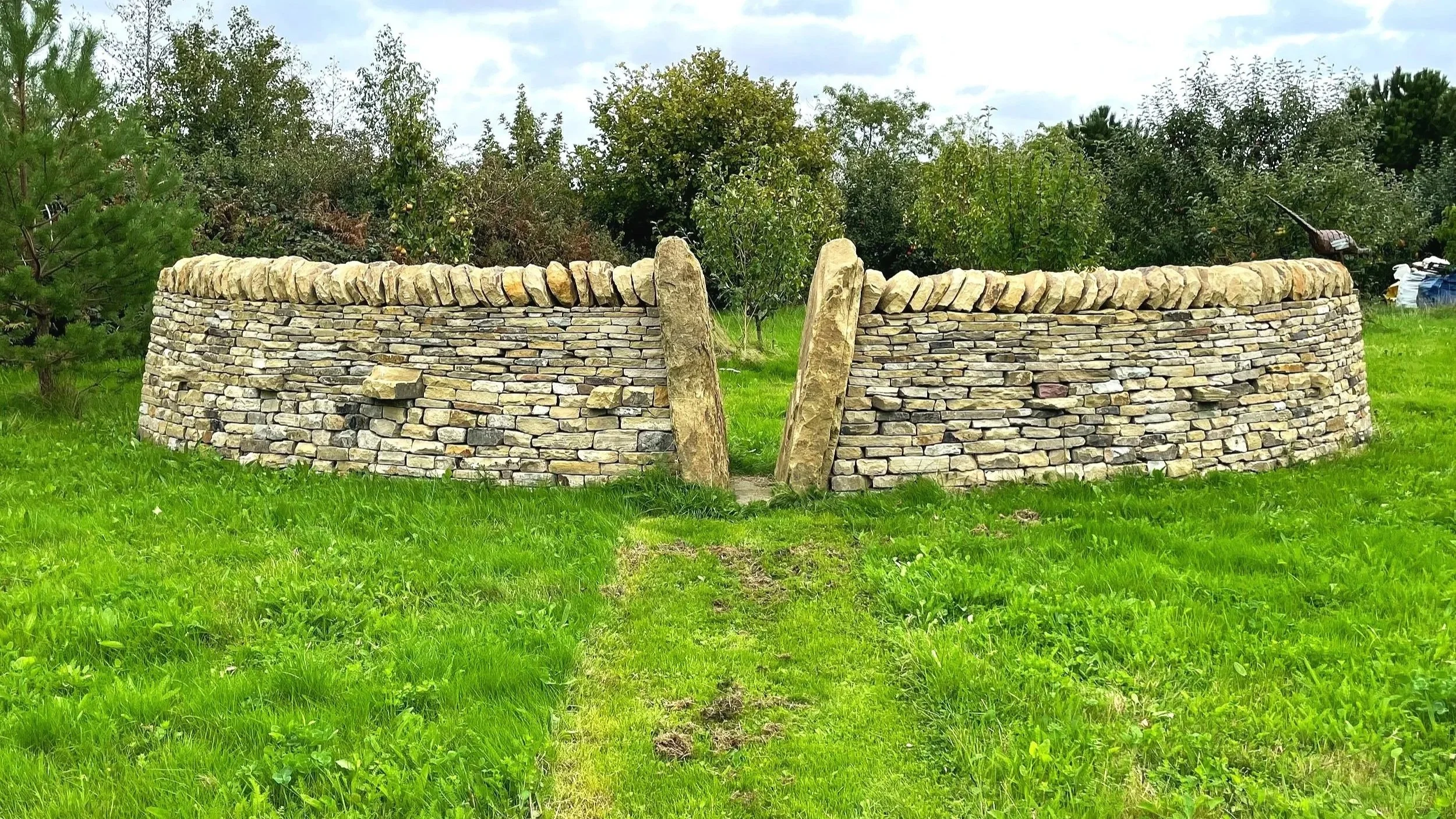 A circular dry stone wall situated on a grassy lawn with small trees and shrubs in the background. The wall is made of stacked flat stones with larger vertical stones at the entrance.
