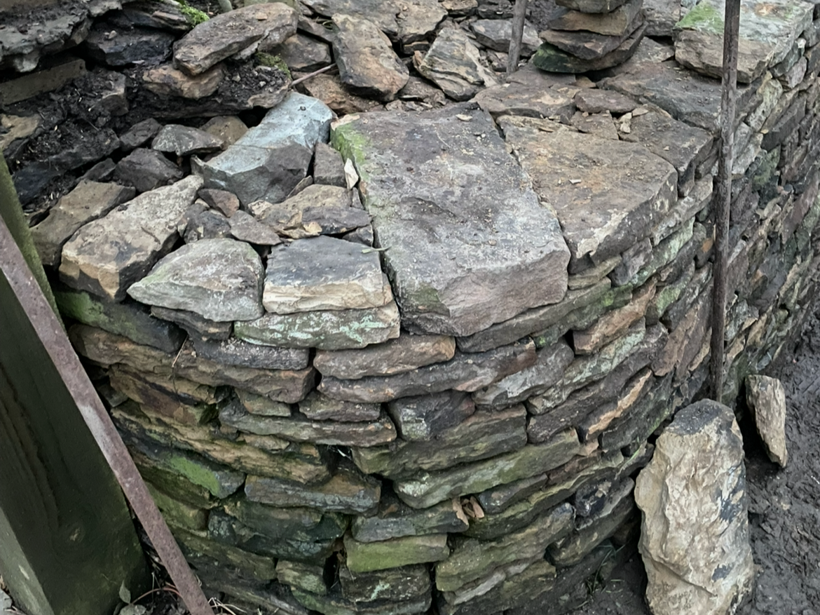 A close-up of a stone wall built with flat, irregularly shaped stones stacked together, with some green moss on the stones.