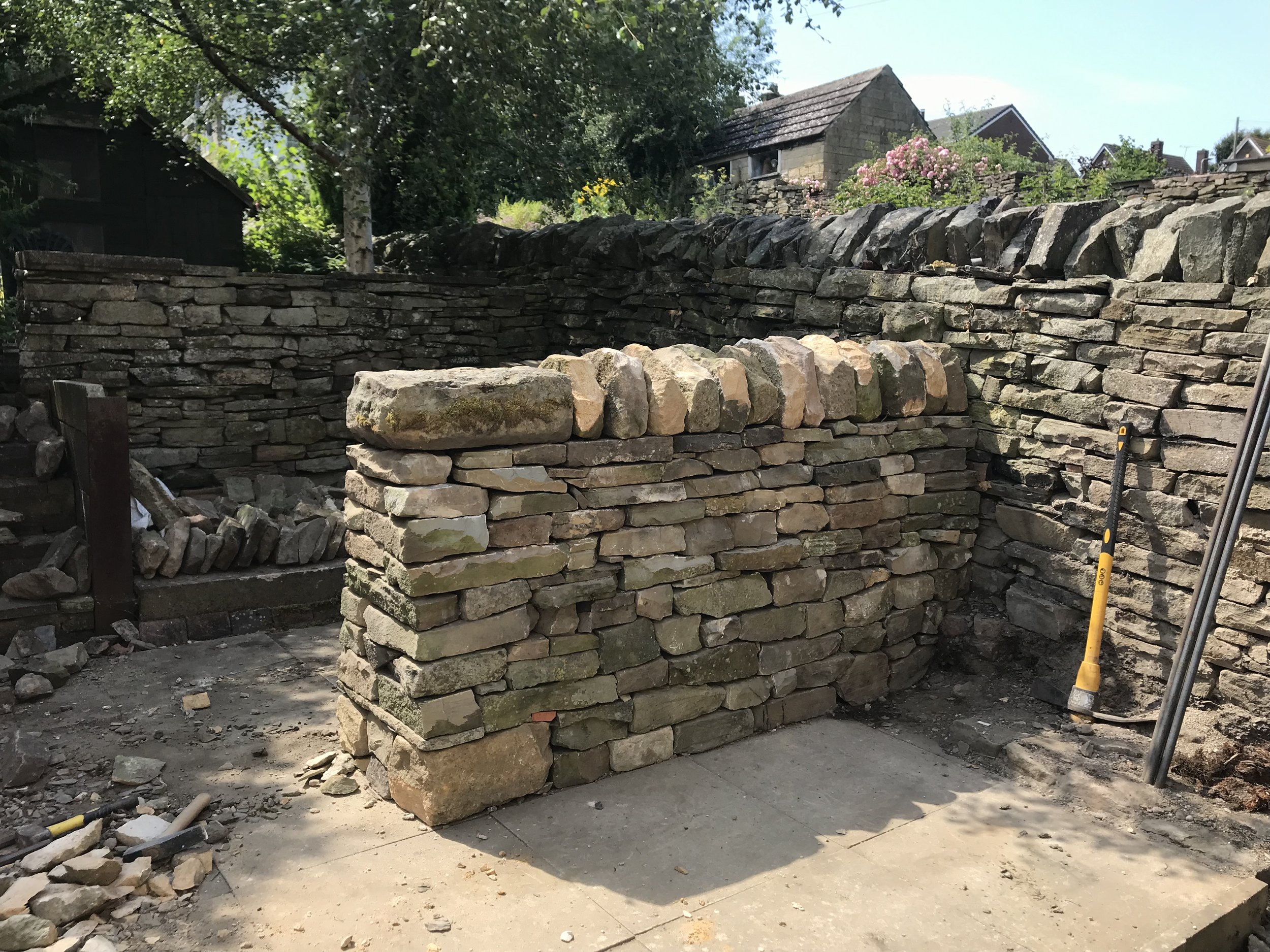 A stone wall under construction with uneven stones and a stack of cut stones nearby, in a shaded outdoor area with trees and houses in the background.