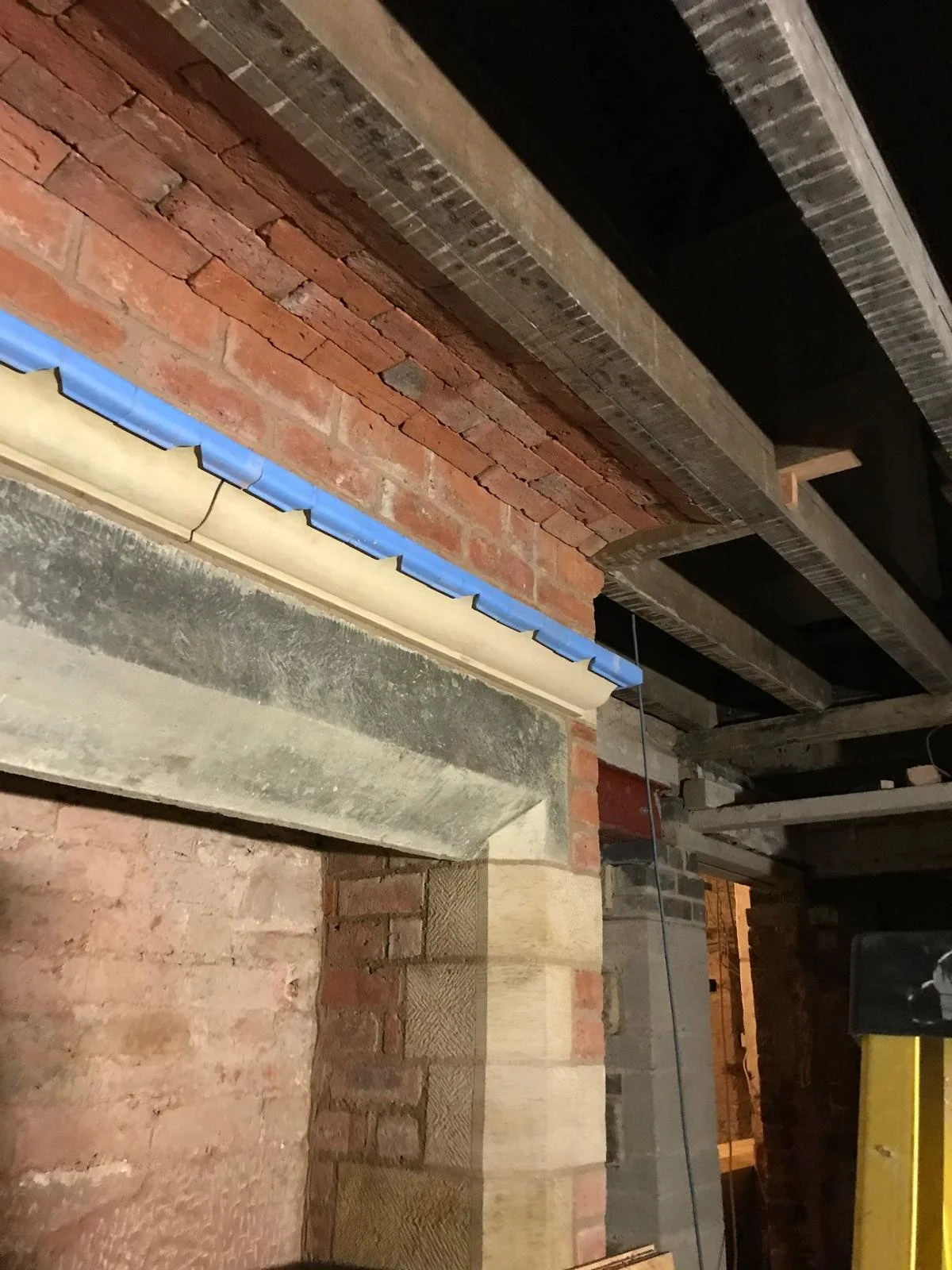 Inside a building under construction with exposed brick walls, concrete beams, and unfinished ceiling with wooden supports and framing.