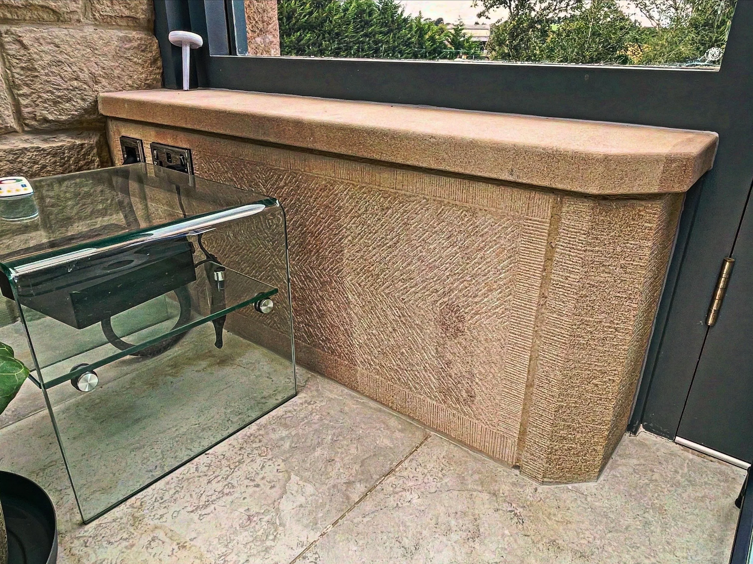 A view of an indoor space showing a stone wall and a large window with outdoor greenery. There's a beige textured stone or wood structure next to the wall, with three electrical outlets on it. A reflective glass table with rounded edges is partially visible in the foreground.