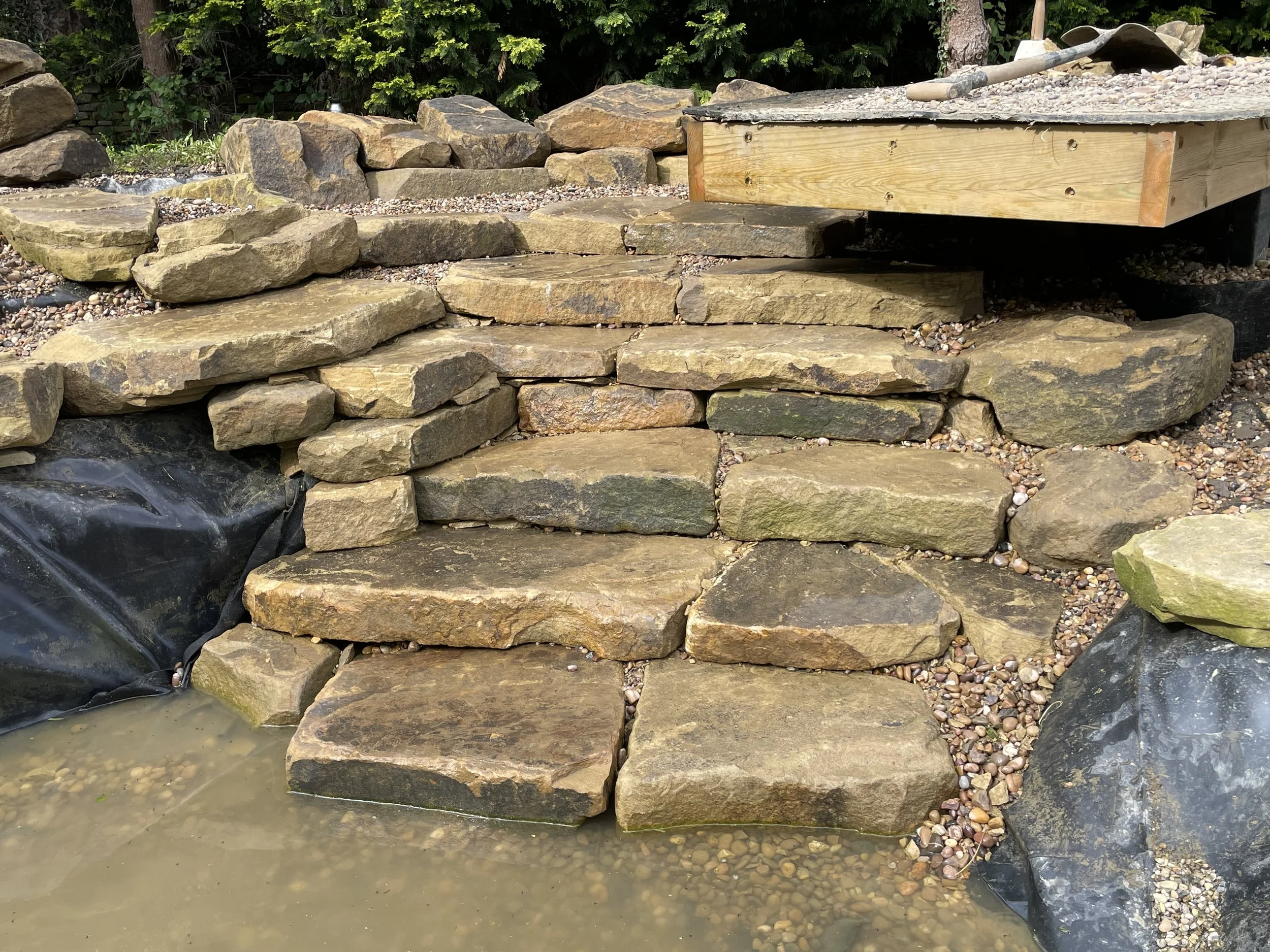 Stacked large and small rocks forming a garden wall with a wooden structure on top, a shovel resting on the structure, and greenery in the background.