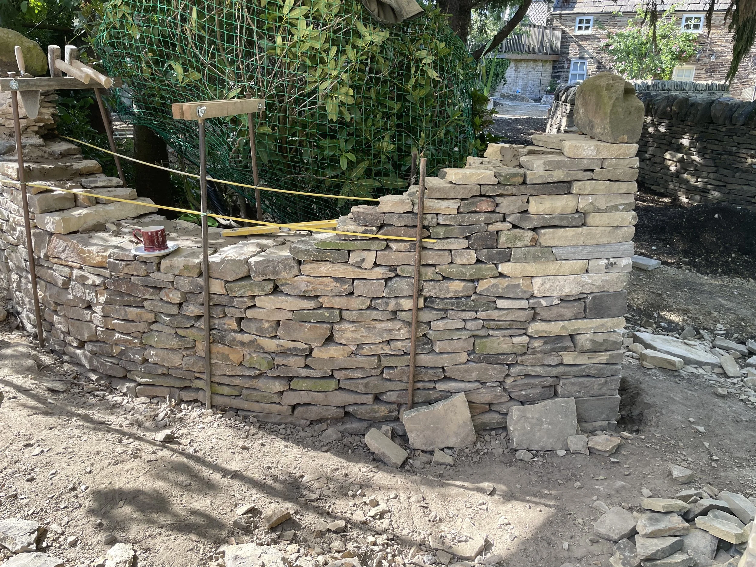 A stone retaining wall under construction with tools attached to it, surrounded by dirt and greenery, and a large stone on top of the wall. In the background are trees and stone buildings.