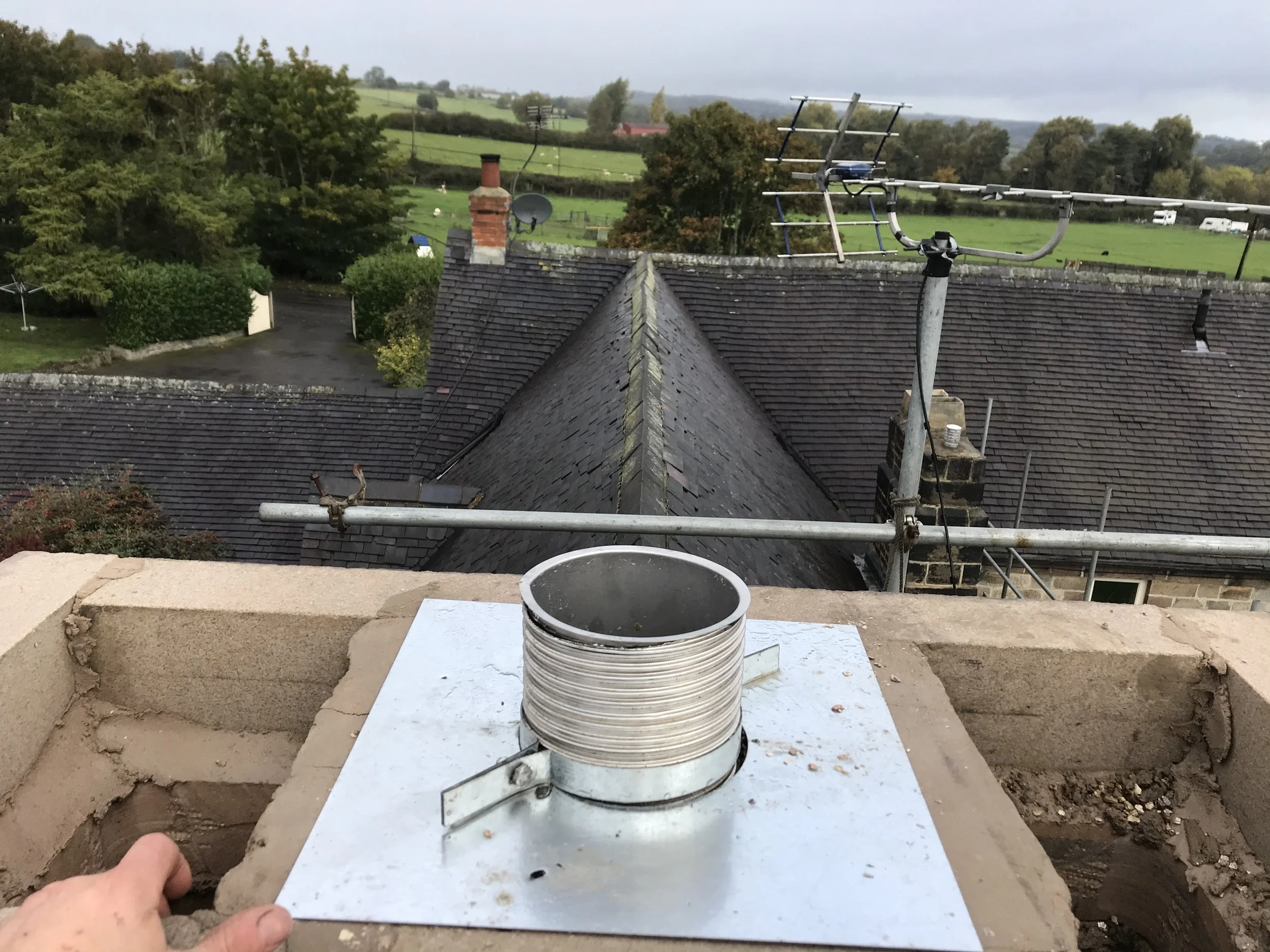 View from a rooftop under construction with a small circular vent pipe in the foreground, weathered shingles on the roof, and a rural landscape with green fields, trees, and distant houses in the background.