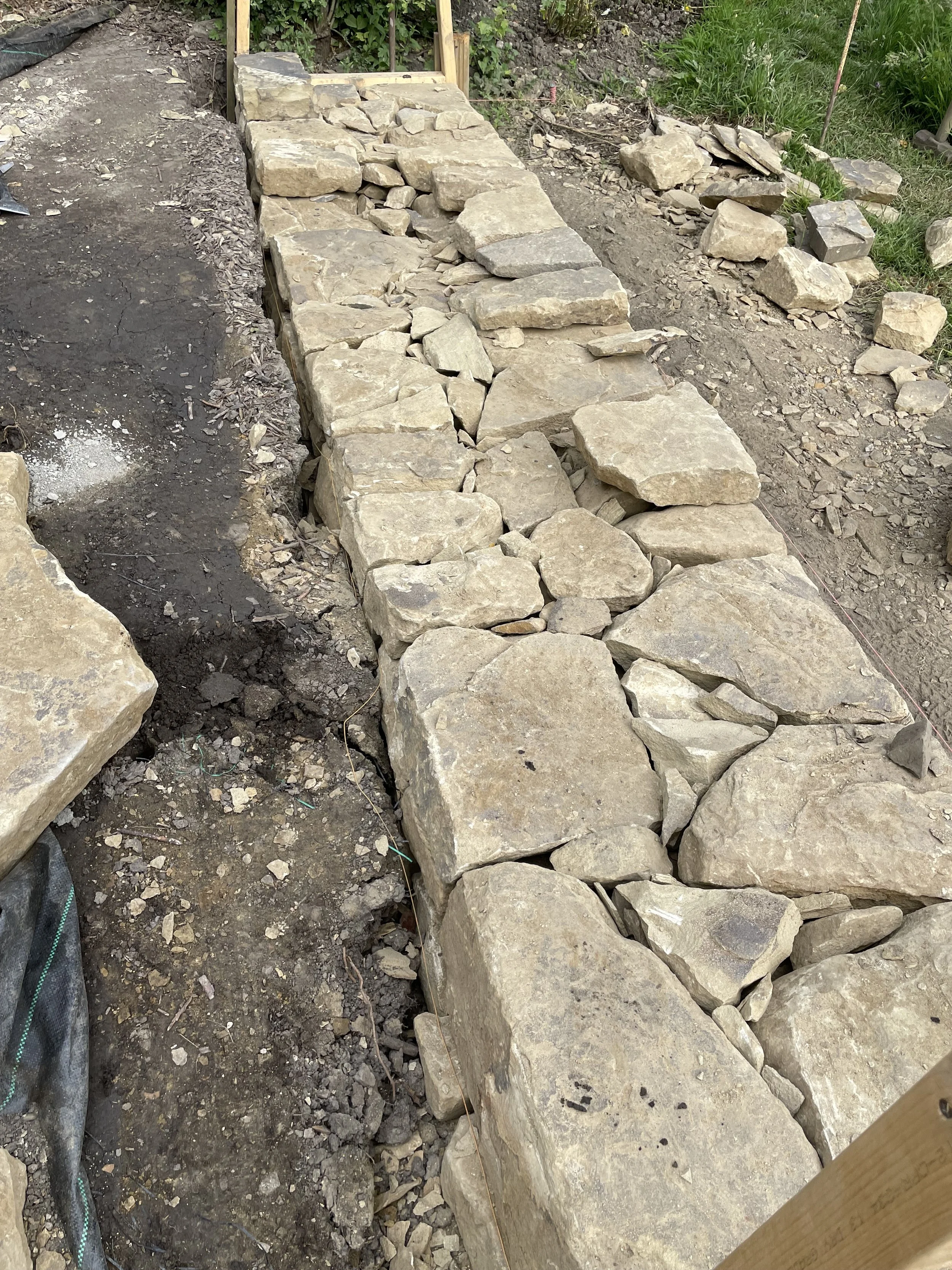 A partially built stone walkway or wall with large uneven rocks arranged in a line on a dirt surface, with some smaller stones scattered nearby. Green foliage is visible in the background.