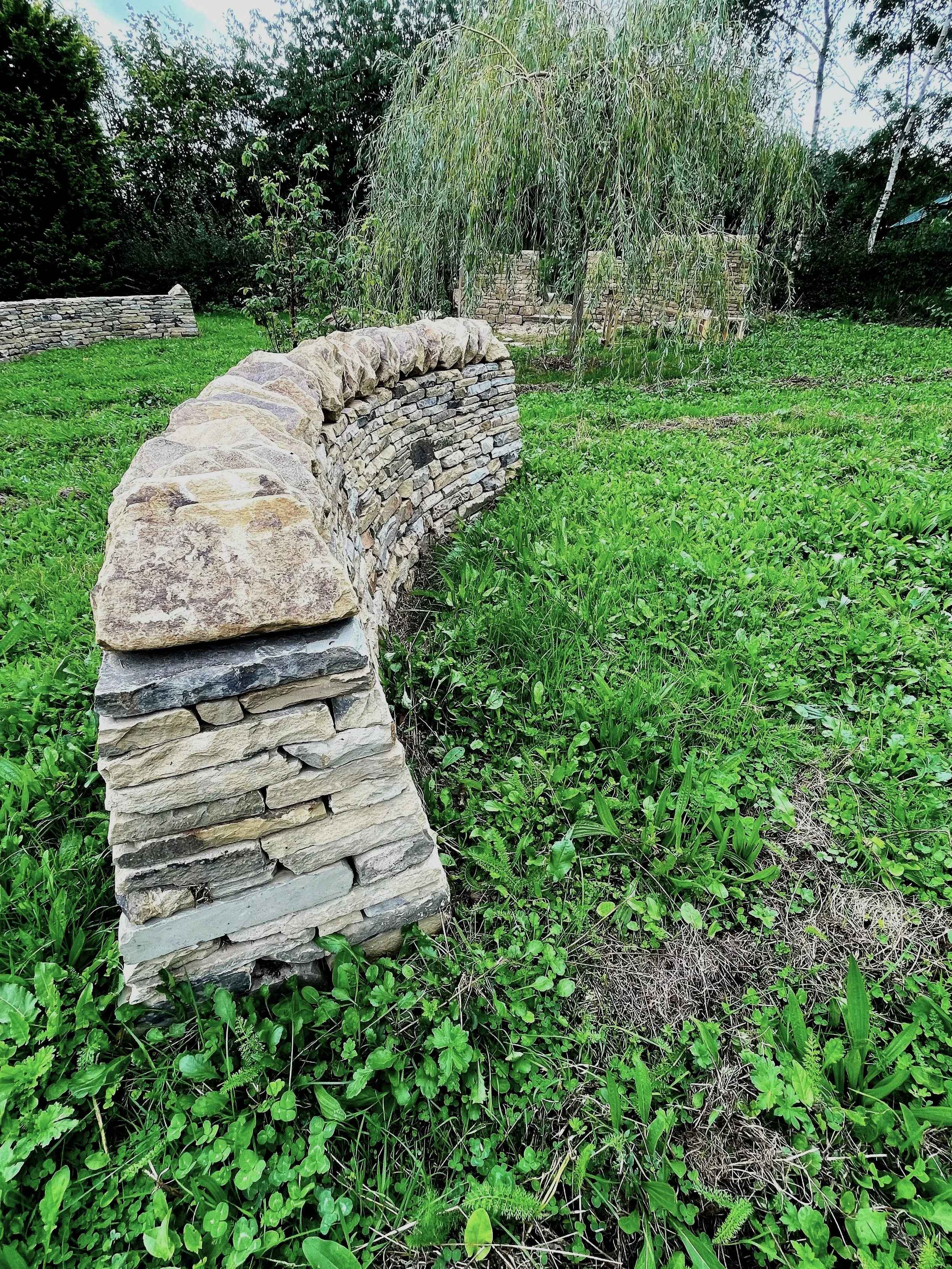 A grassy outdoor area with a stone wall and arch, a weeping willow tree, and surrounding green trees and plants.