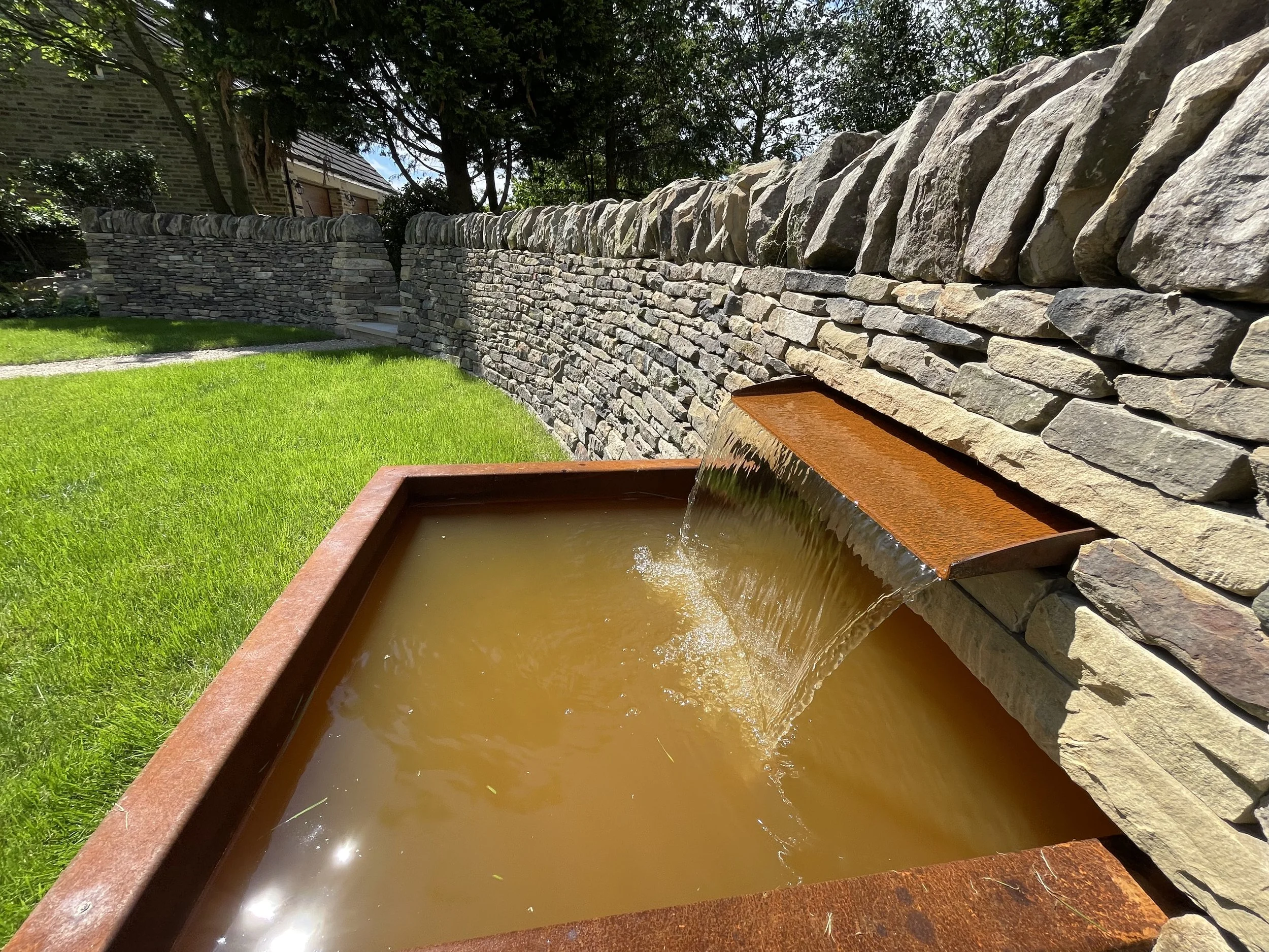 A stone retaining wall with a concrete cap, a small staircase, and a pond with a rusty metal edge in a lush green yard with trees and a house in the background.