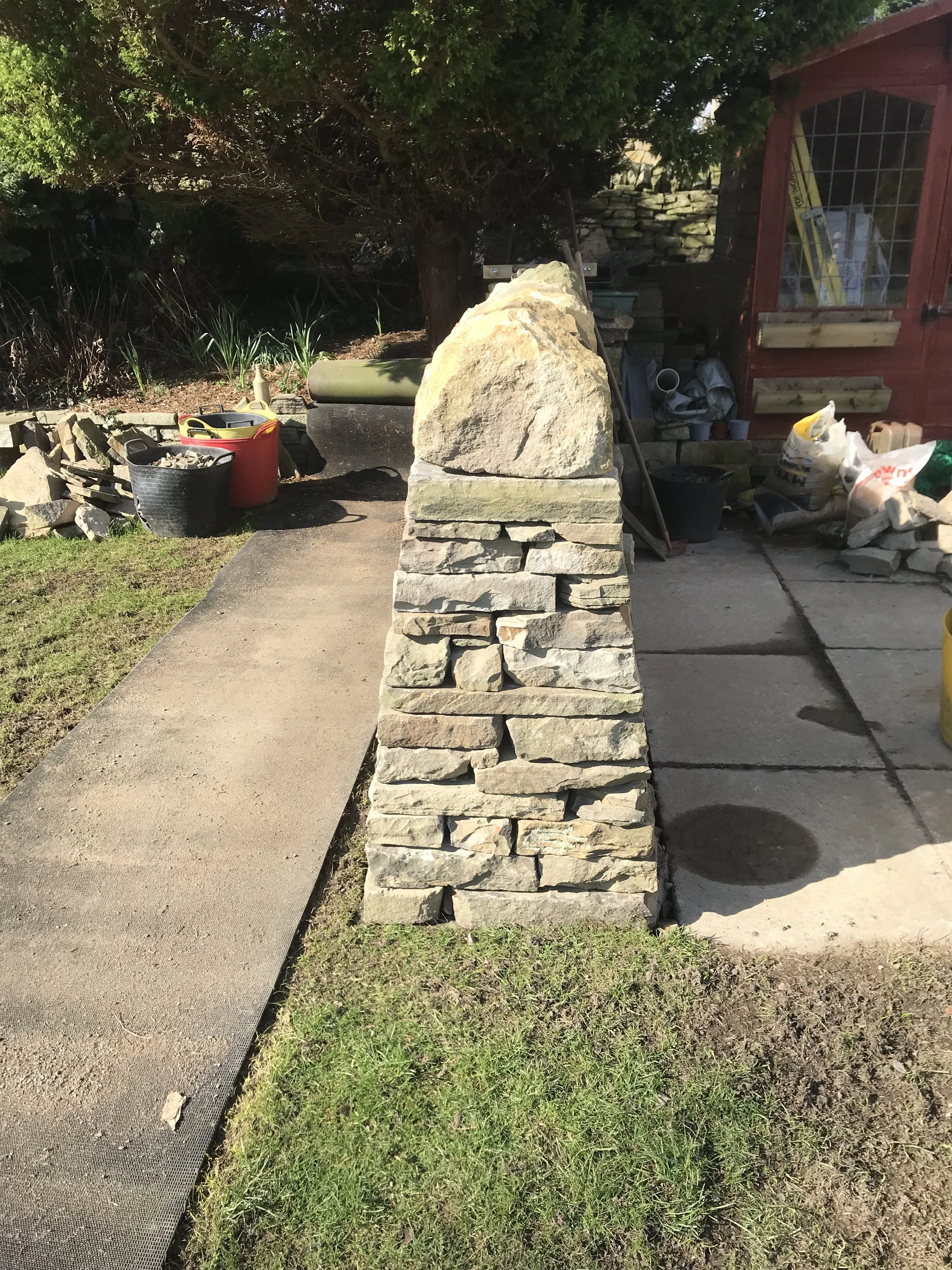 Side view of a landscaped garden with a stone wall and a small red shed, featuring gardening supplies and tools, with plants and trees in the background.