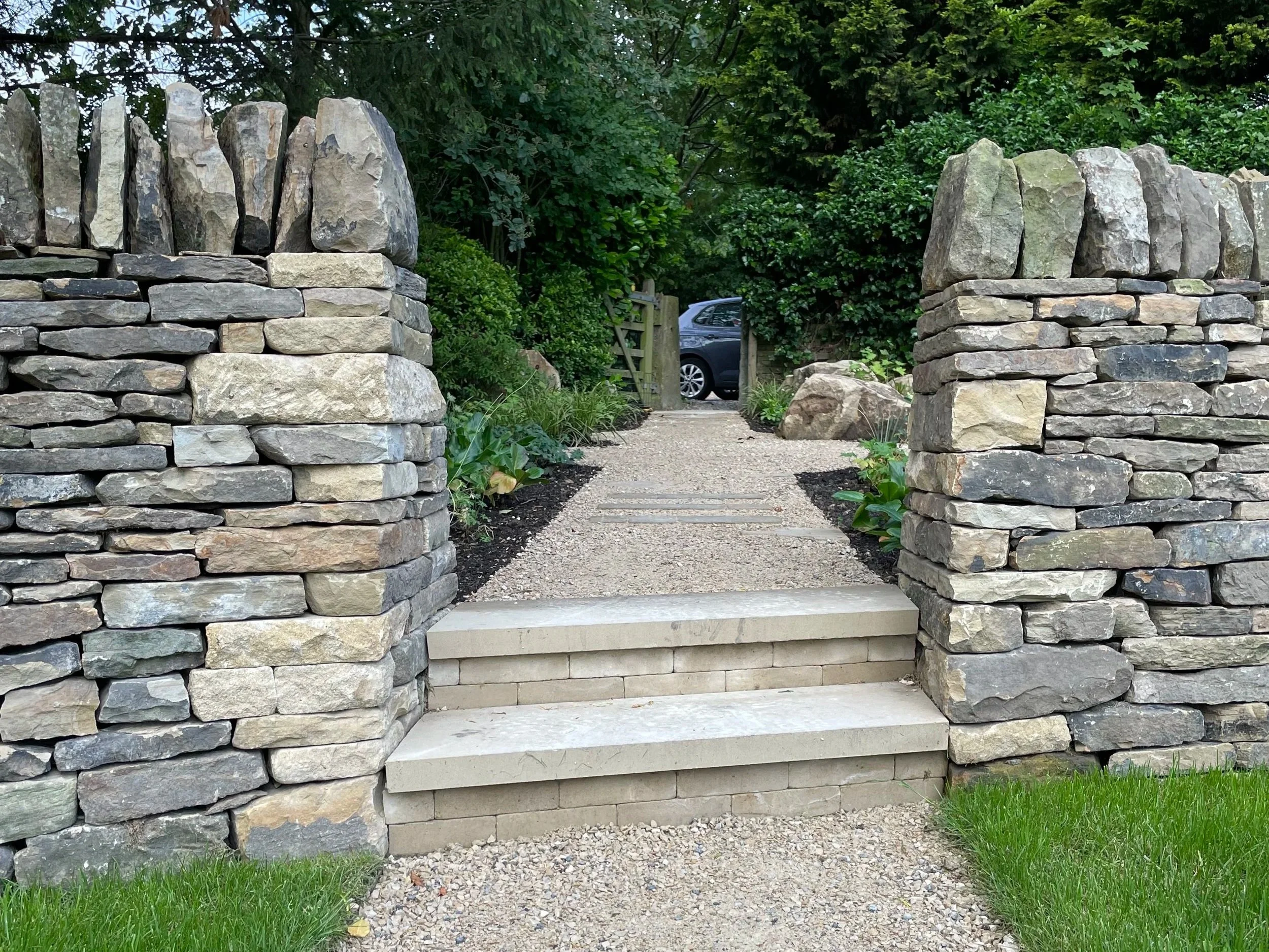 A stone pathway with steps flanked by stone walls leading to a metal gate, with greenery and trees in the background.