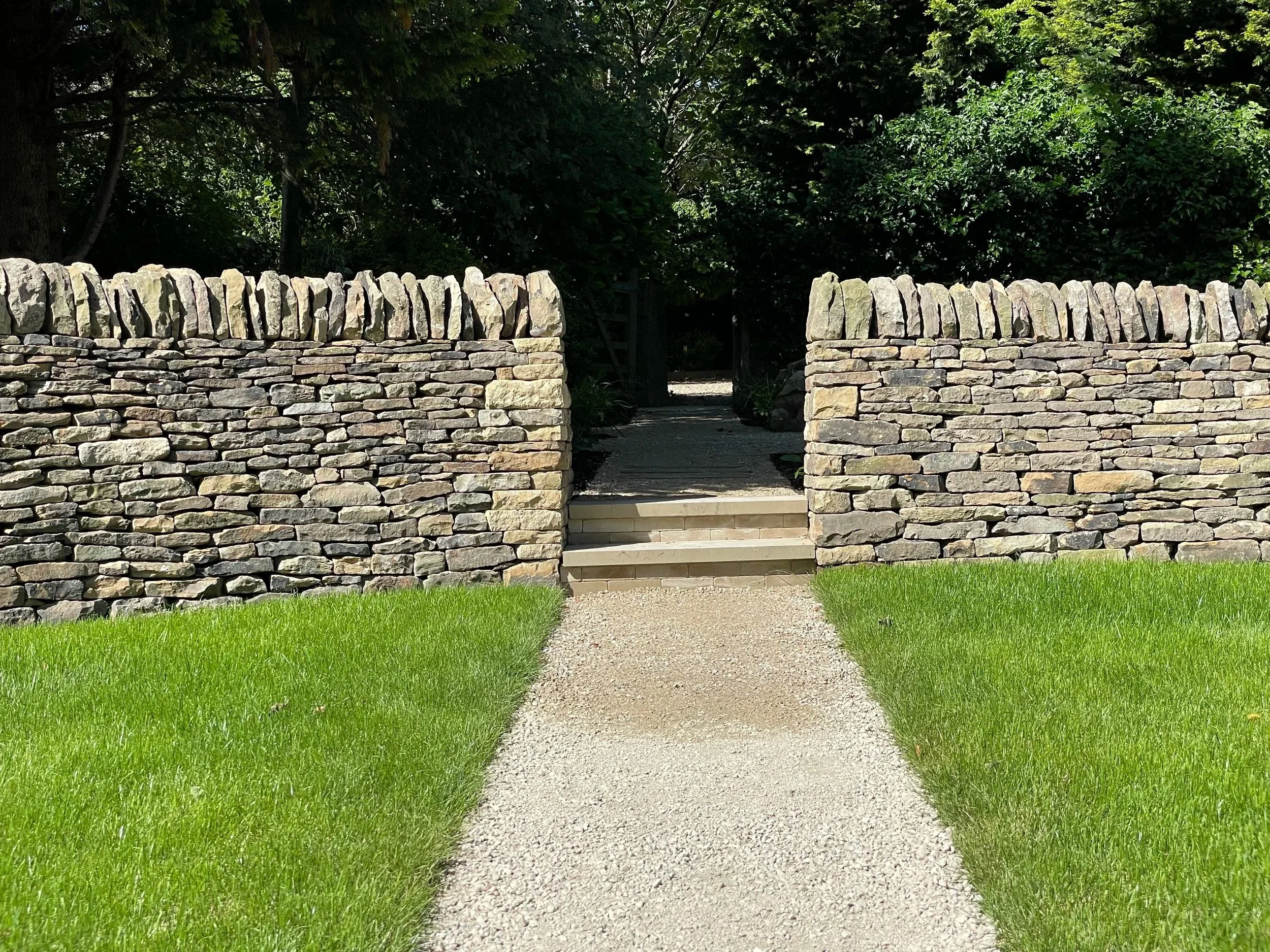 A stone pathway leads through a garden with a stone wall on either side. The path is bordered by green grass, and there are trees and dense foliage in the background.