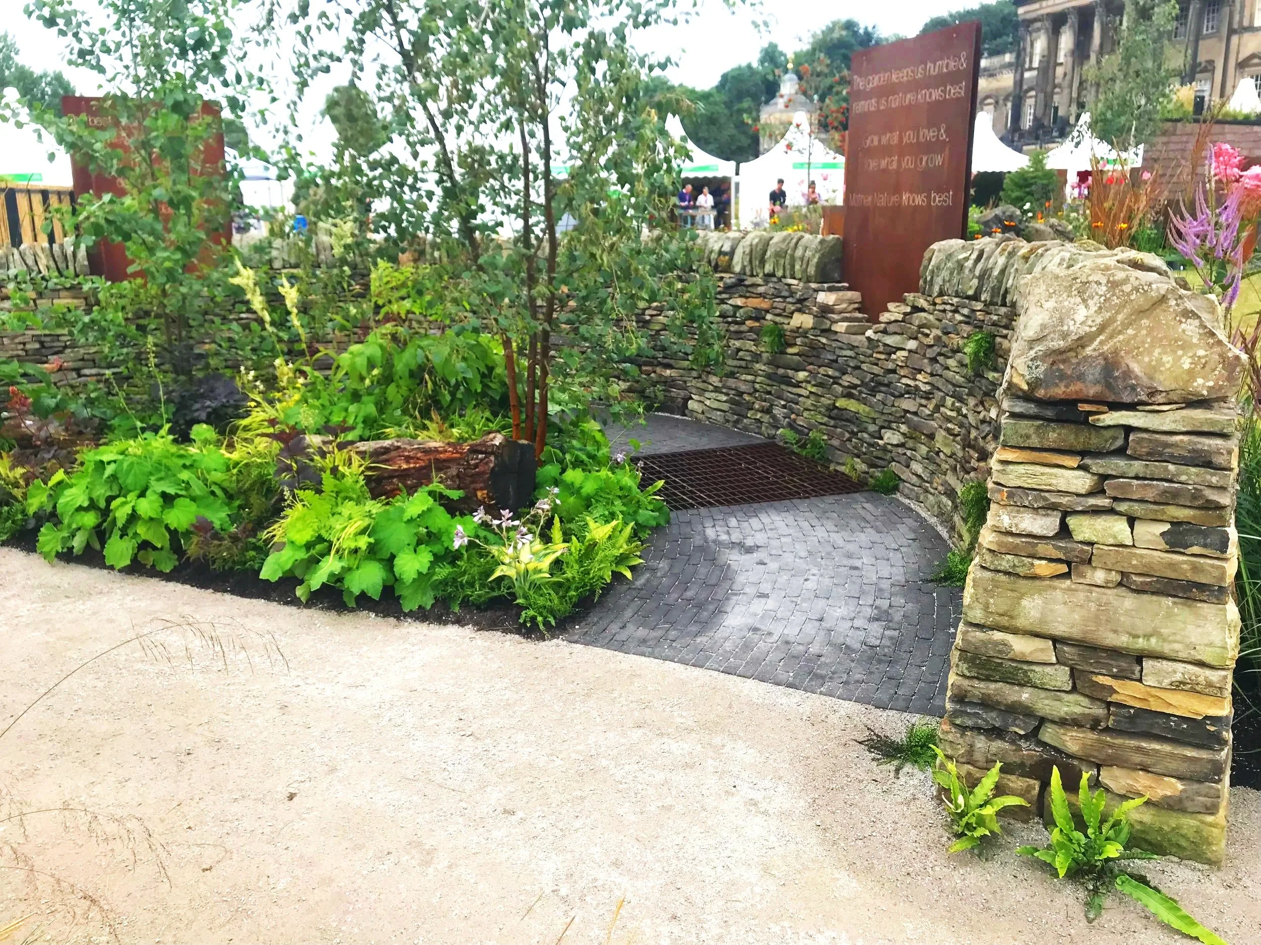 A decorative stone wall with a metal grate below, surrounded by vibrant green plants and flowers, with tents and people in the background at an outdoor event.