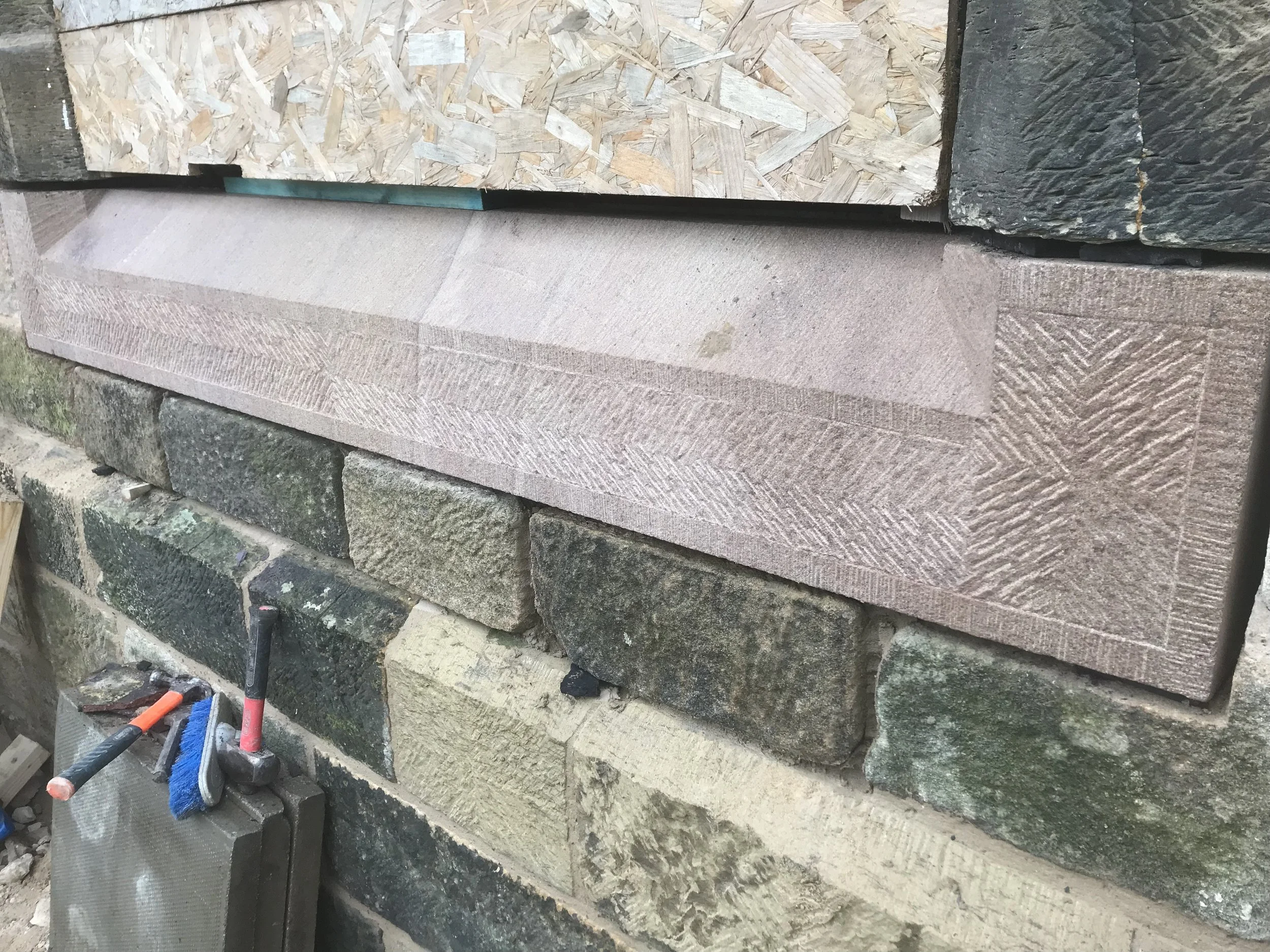 Construction site with a wall built from different types of stone and brick, including a section of OSB wood paneling, mortar, and various construction tools.