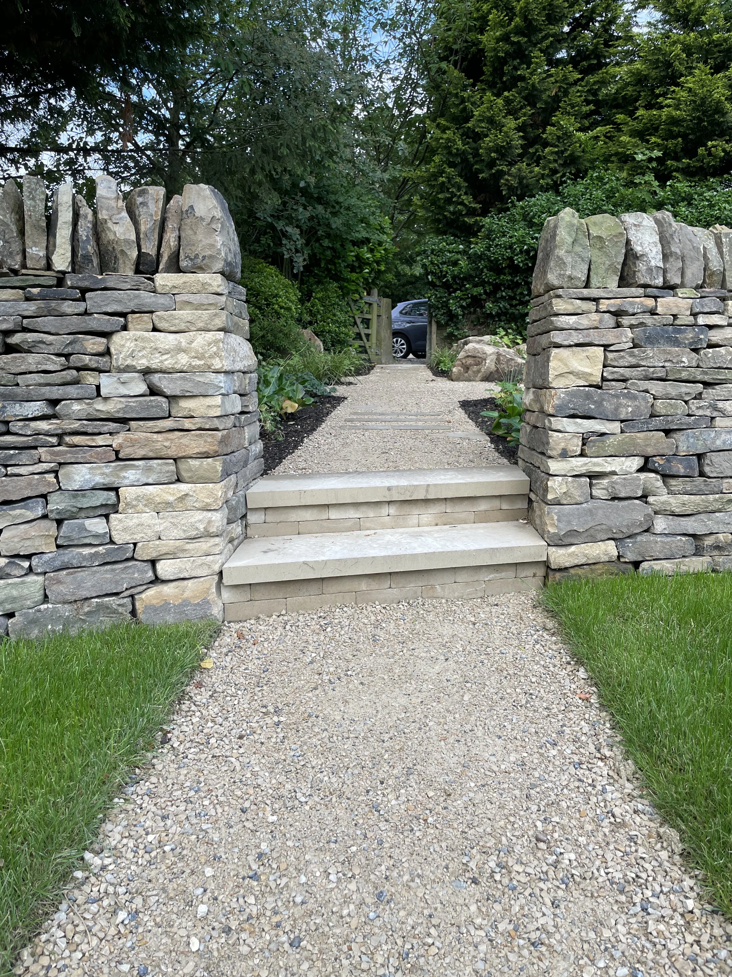 A pathway with small stones leading through a garden gate, flanked by stone walls and surrounded by green trees and bushes.