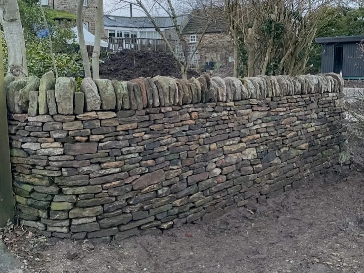 A dry stone retaining wall made of flat, irregular stones, with larger stones on top. Behind the wall are trees, bushes, and a backyard with buildings and a shed.