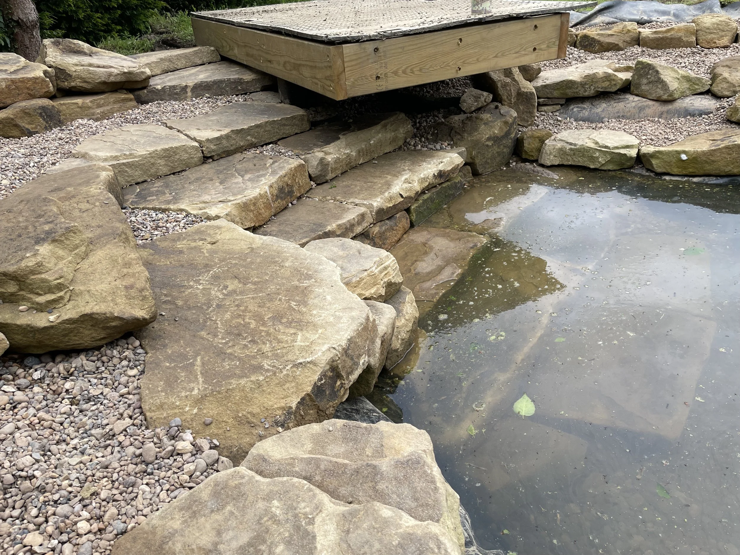 A lakeside scene with large flat rocks and smaller gravel stones along the shore, a wooden platform over the water, and some plants and trees in the background.
