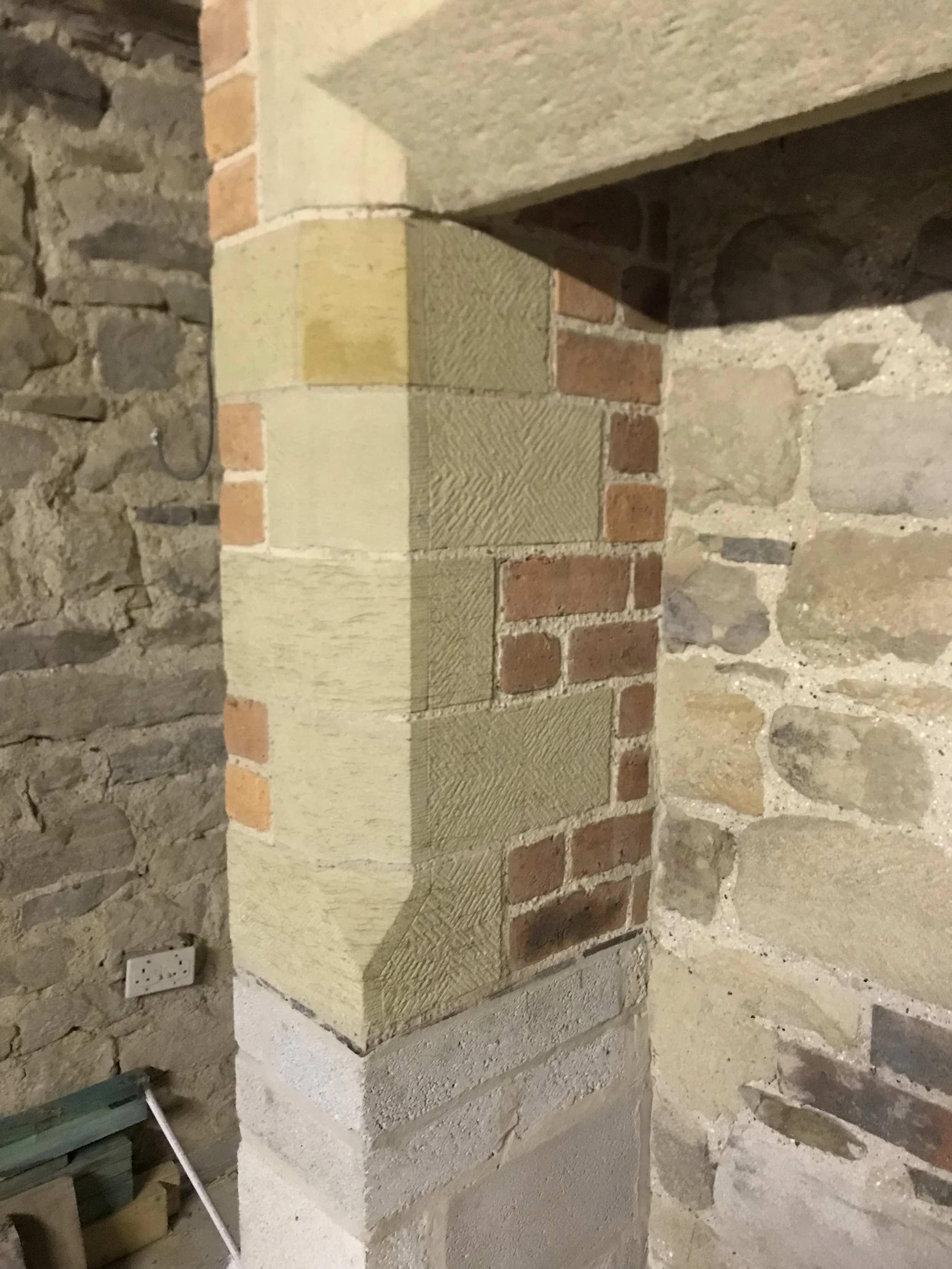Close-up of a construction or renovation site showing a brick and stone wall with a newly built or repaired section. The section features a combination of bricks, bricks covered with plaster, and stonework on the surrounding walls.