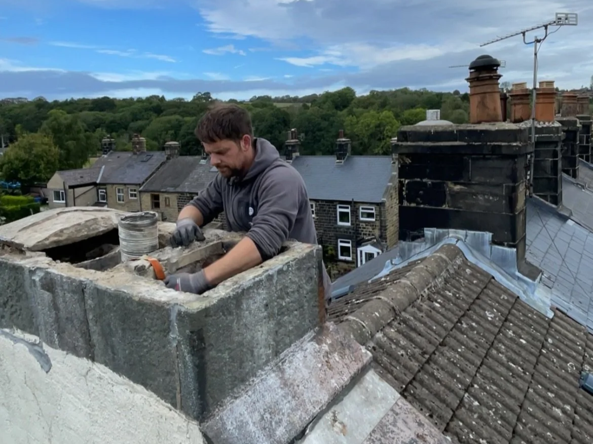 Man working on a chimney repair on the roof of a house with a background of trees and adjacent rooftops.