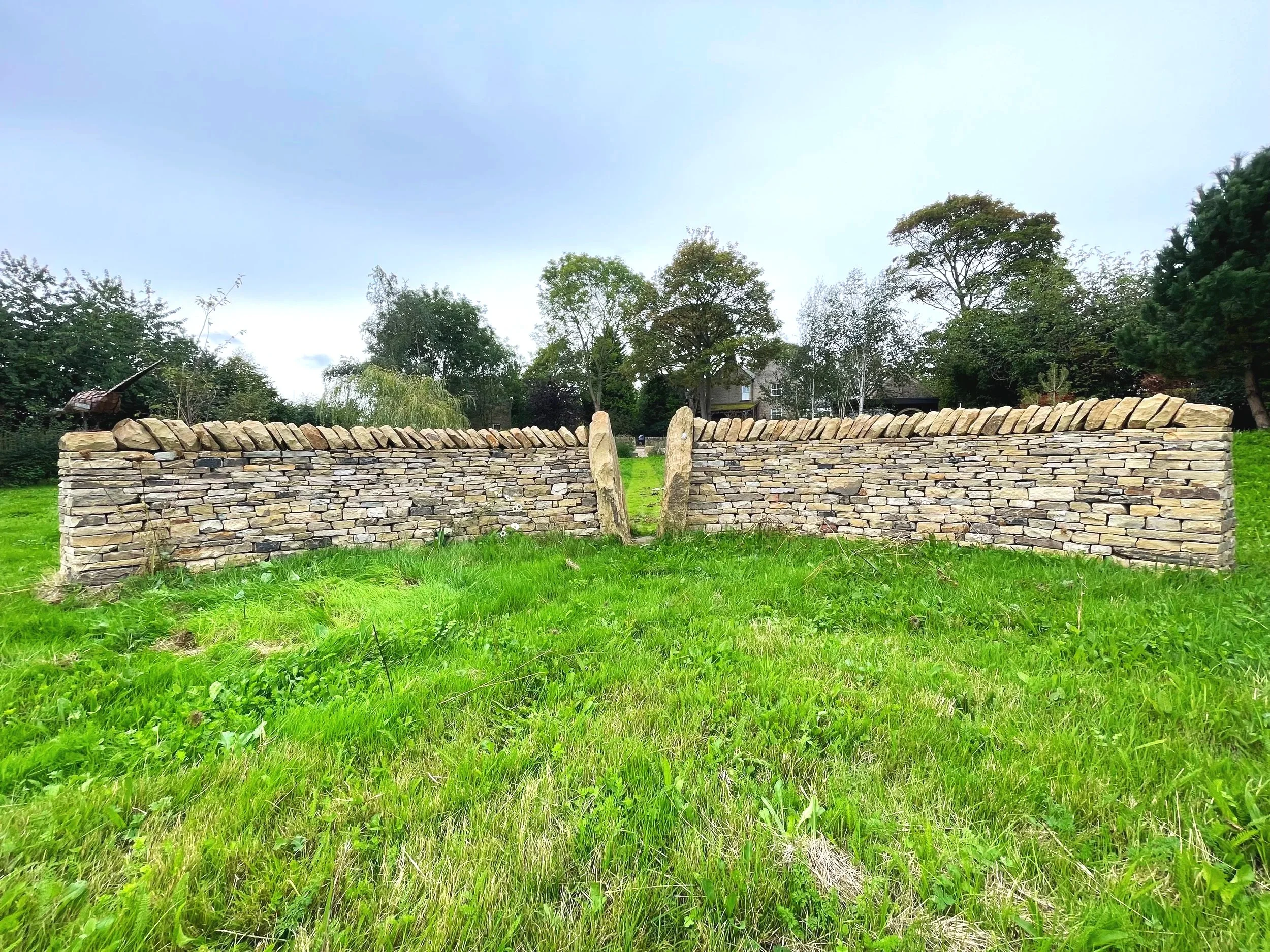 A stone wall in a grassy field with trees in the background and a house visible behind the trees.