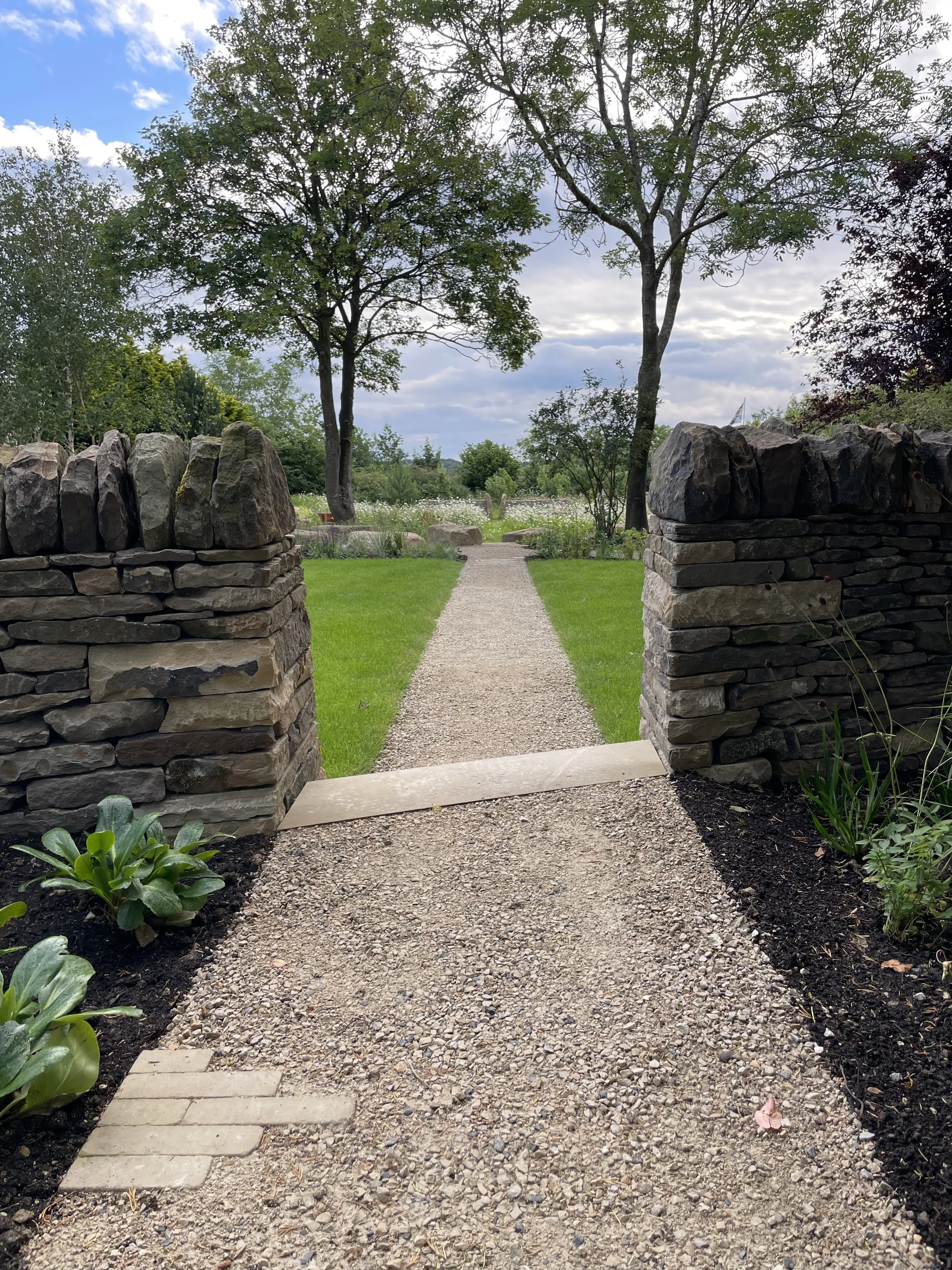A garden pathway made of small gravel stones flanked by green plants and flower beds, bordered by stone pillars leading to a park with trees and a partly cloudy sky.