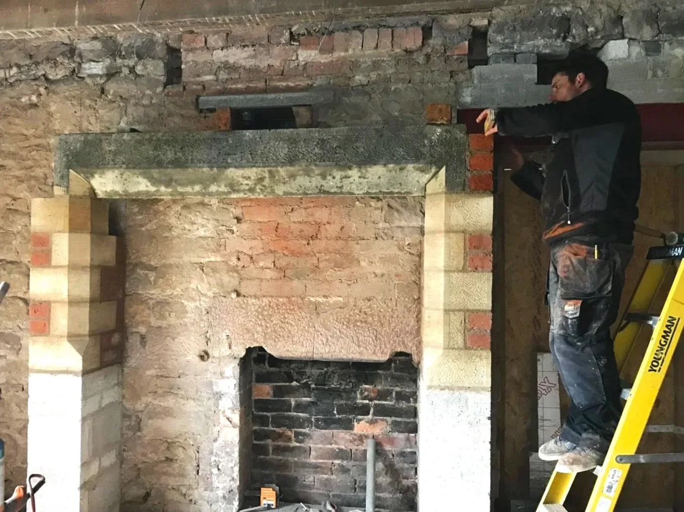 A man standing on a yellow ladder working on a brick fireplace in a room under renovation.