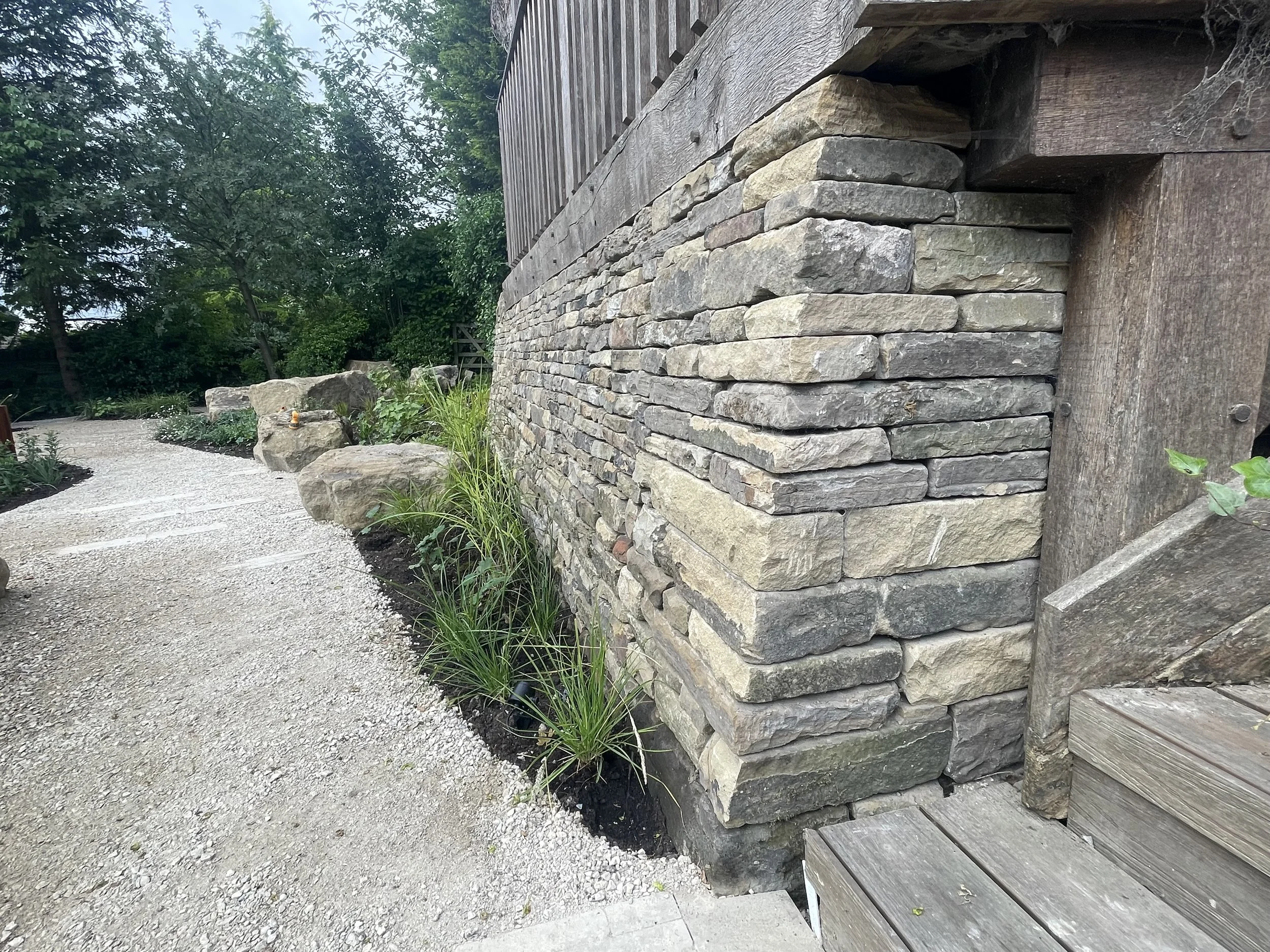 A stone and wood exterior wall of a building with greenery and a gravel pathway in front.