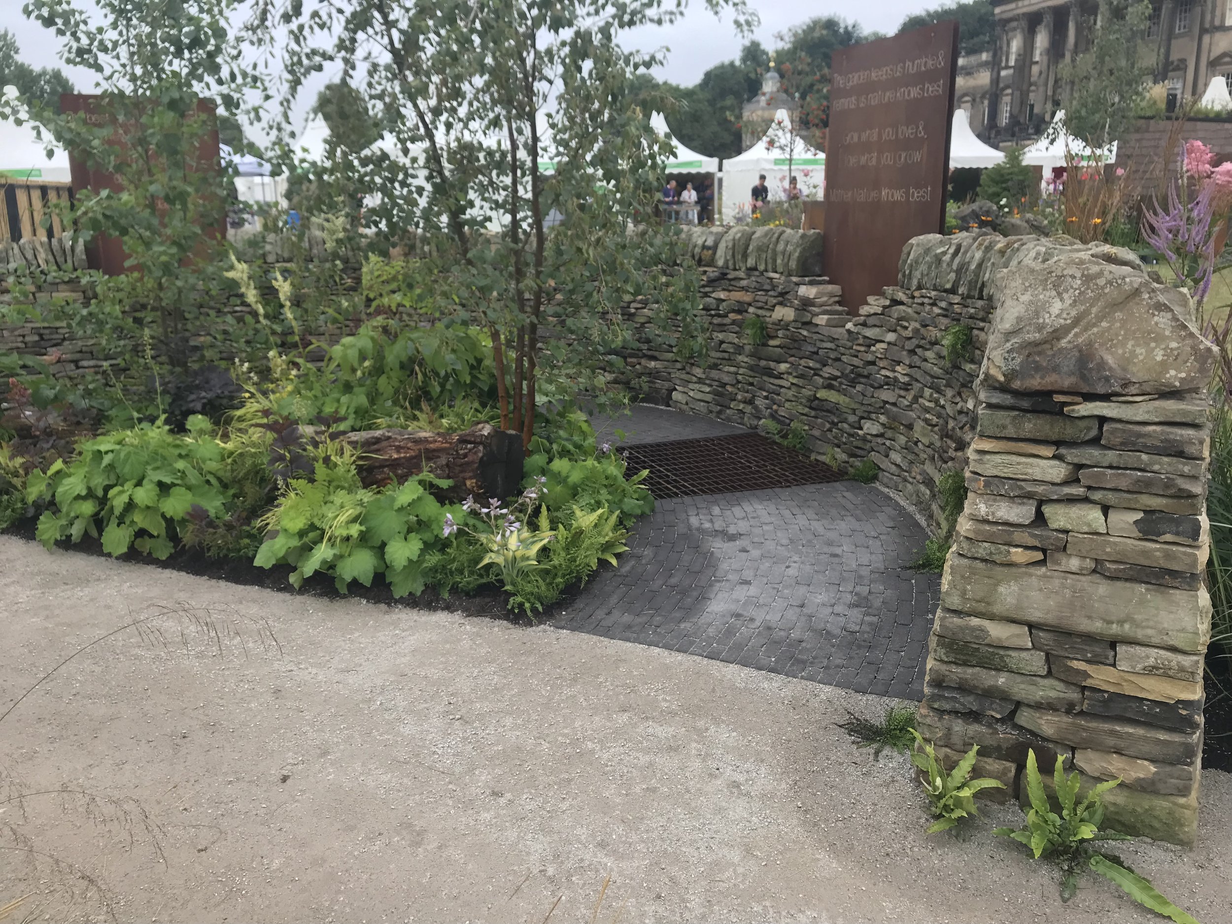 A garden area enclosed by a stone wall with lush green plants and flowers. There is a pathway in the foreground and tents with people in the background, suggesting an outdoor event. A large sign with words about nature is visible behind the wall.