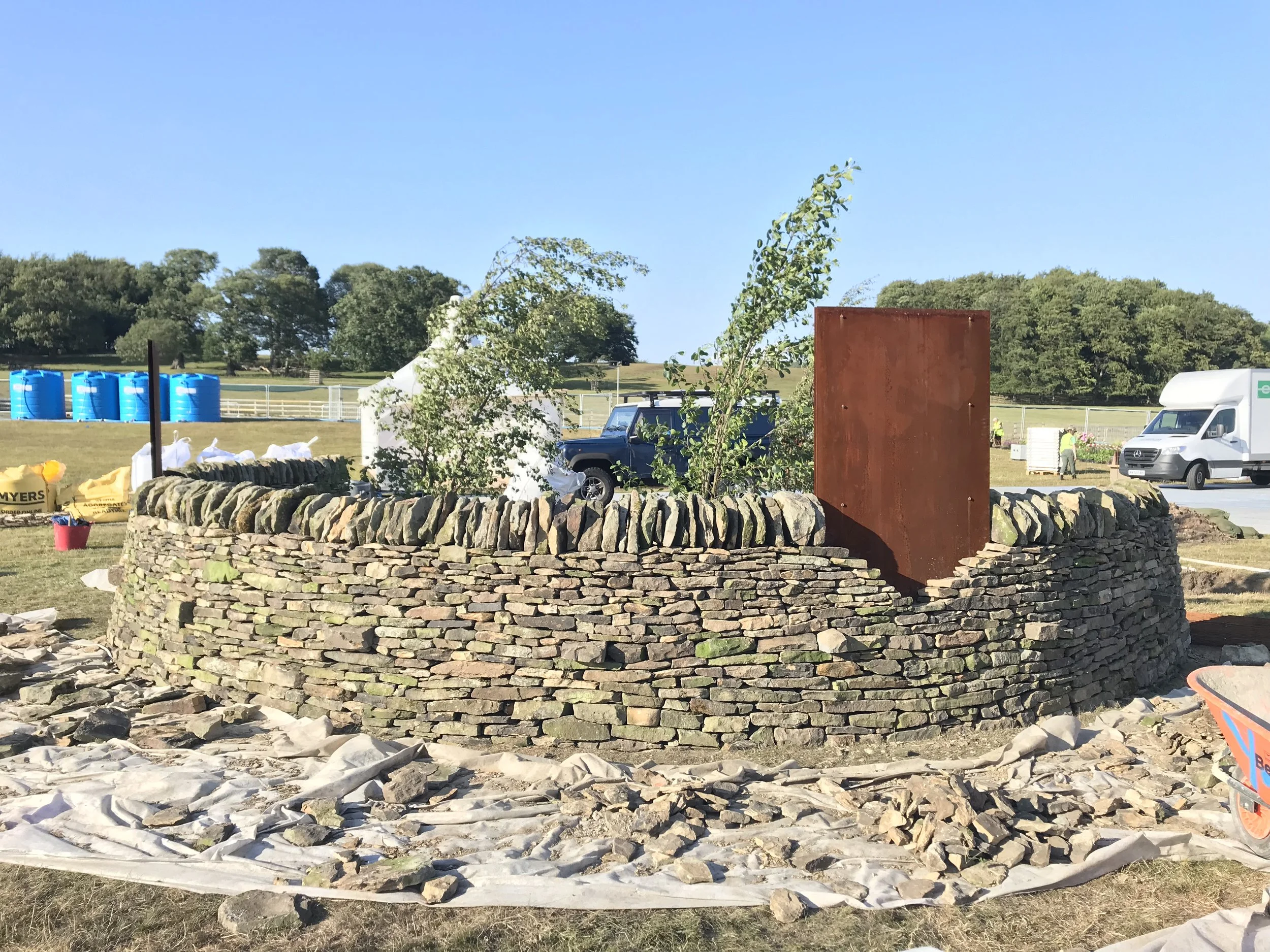 A circular stone wall under construction outdoors with a rusty metal sheet in the middle. There are trees, vehicles, and blue water tanks in the background, along with construction materials and bags of supplies on the ground.