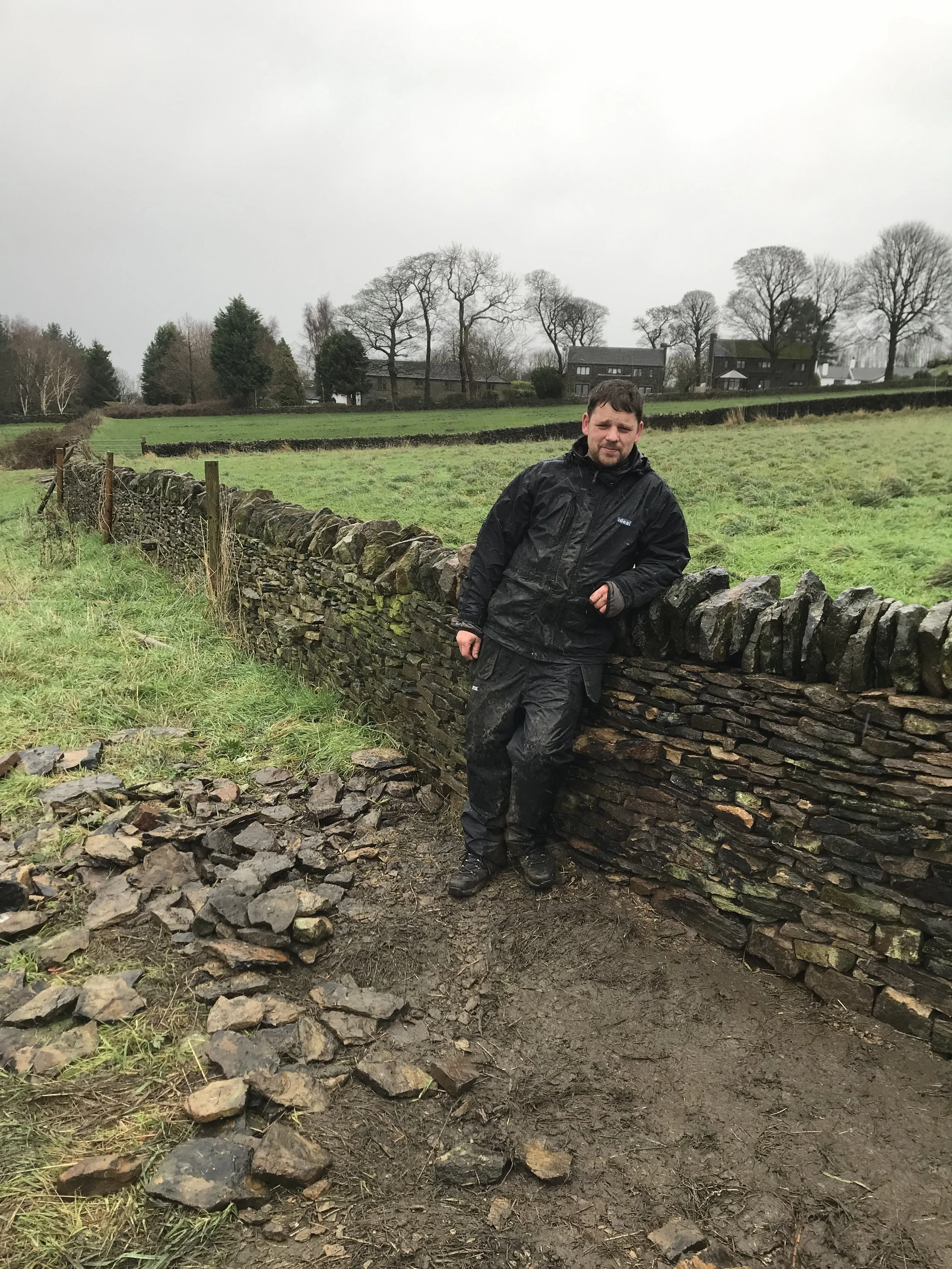 A man in black waterproof outdoor clothing standing beside a large, stacked dry stone wall in a rural area with green fields, trees, and houses in the background on a cloudy day.