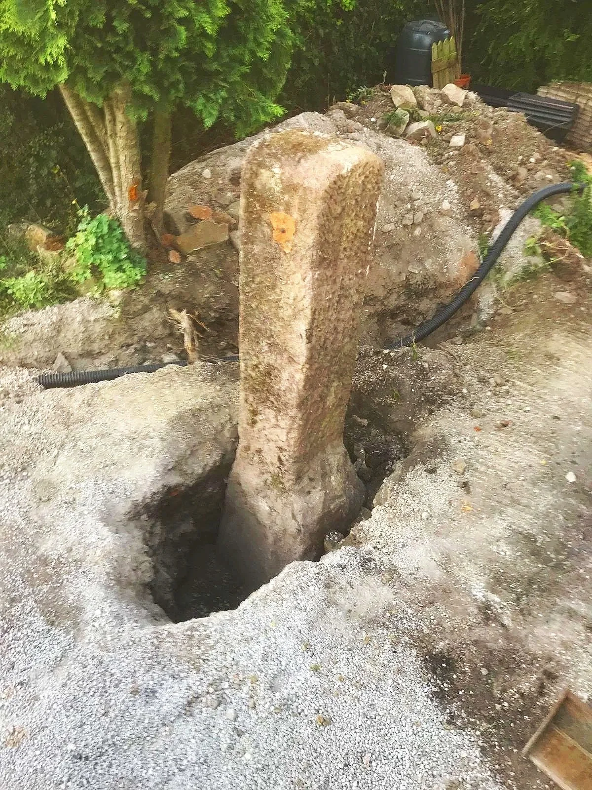 A large stone pillar is being removed from the ground using heavy machinery, with a deep hole and loose dirt around it. There are trees, plants, and construction materials nearby, including black pipes and a plastic container.