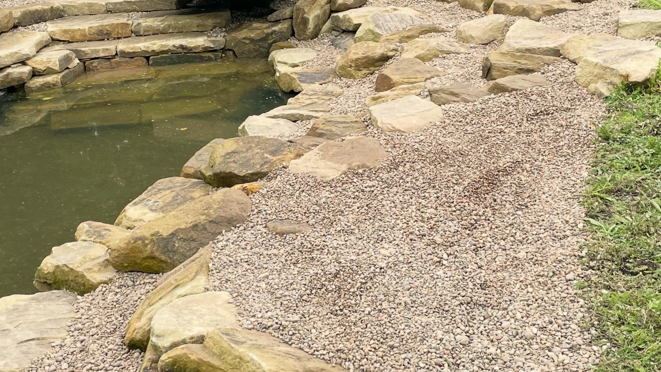 A small pond or water feature bordered by large flat stones and a gravel pathway with some greenery on the side.
