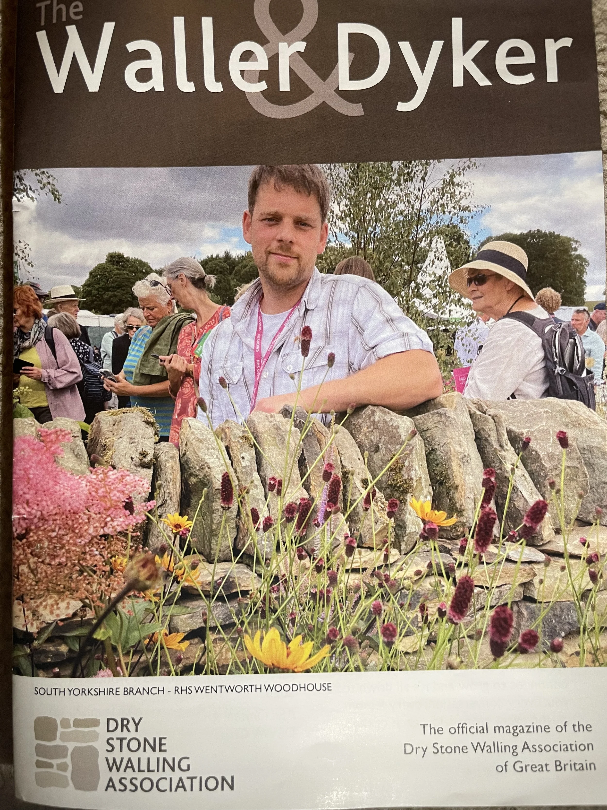 Magazine cover displaying a man with a beard and short brown hair smiling outdoors at a festival with other people, trees, and a cloudy sky in the background. Front of the magazine shows colorful flowers and a stone wall. The magazine is published by the Dry Stone Walling Association of Great Britain, titled 'The Waller & Dyker', and mentions the South Yorkshire Branch and RHS Wentworth Woodhouse.
