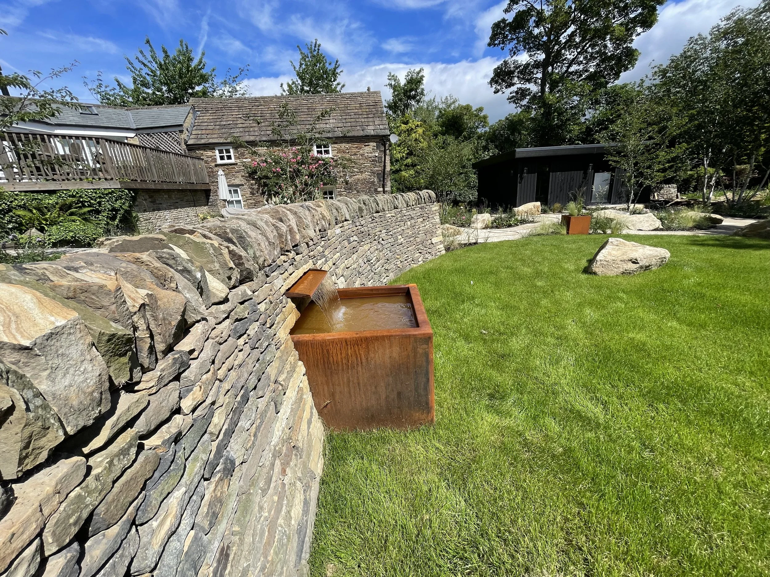A garden with a stone wall, a waterfall feature, green grass, trees, and a house with a balcony and small windows in the background.