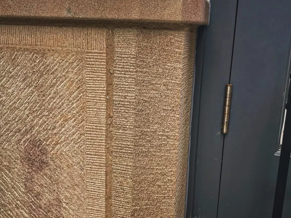 Close-up of textured beige brick wall next to dark gray painted door with a brass hinge.