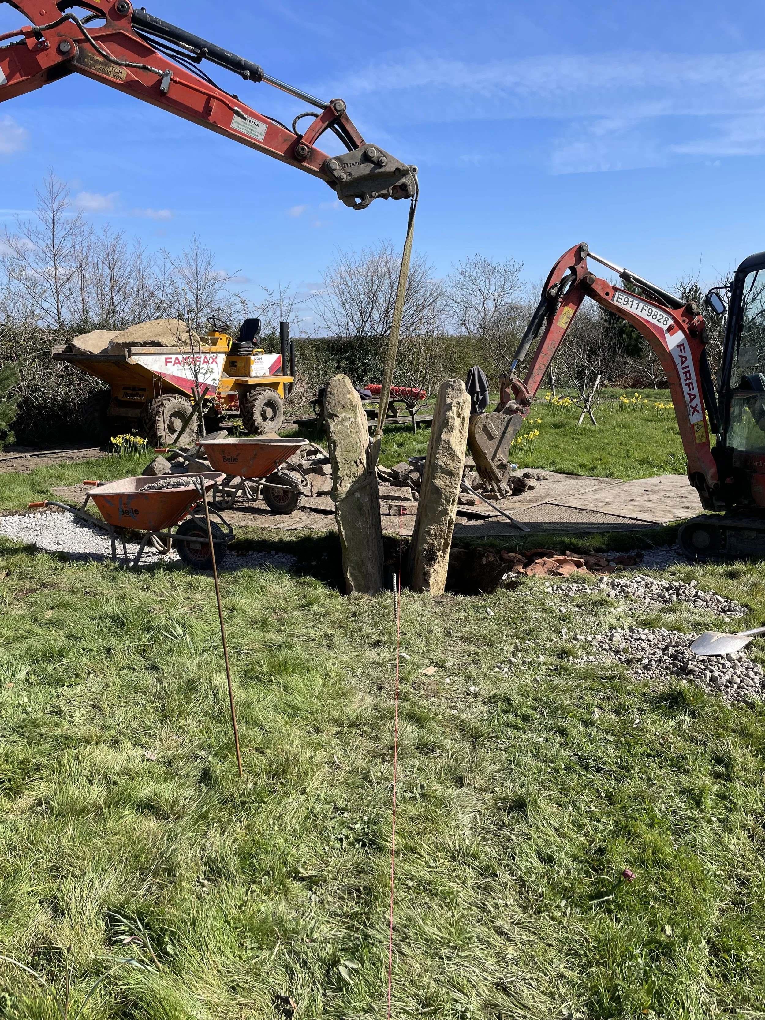 Construction site with excavators, large rocks, wheelbarrows, and a grassy area with yellow flowers under a blue sky with clouds.