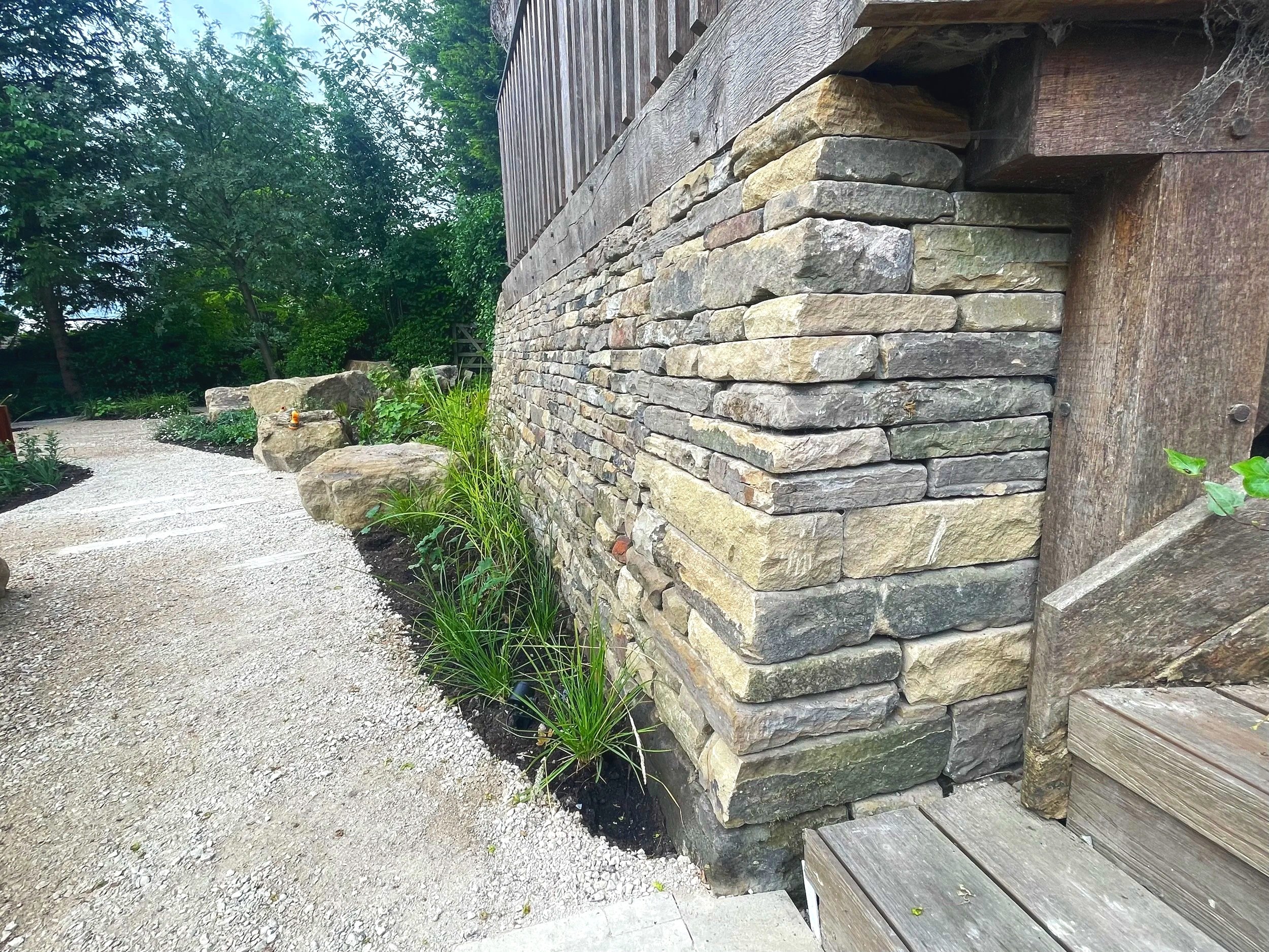 A corner of a stone and wood building with a gravel pathway beside it, surrounded by greenery and trees.