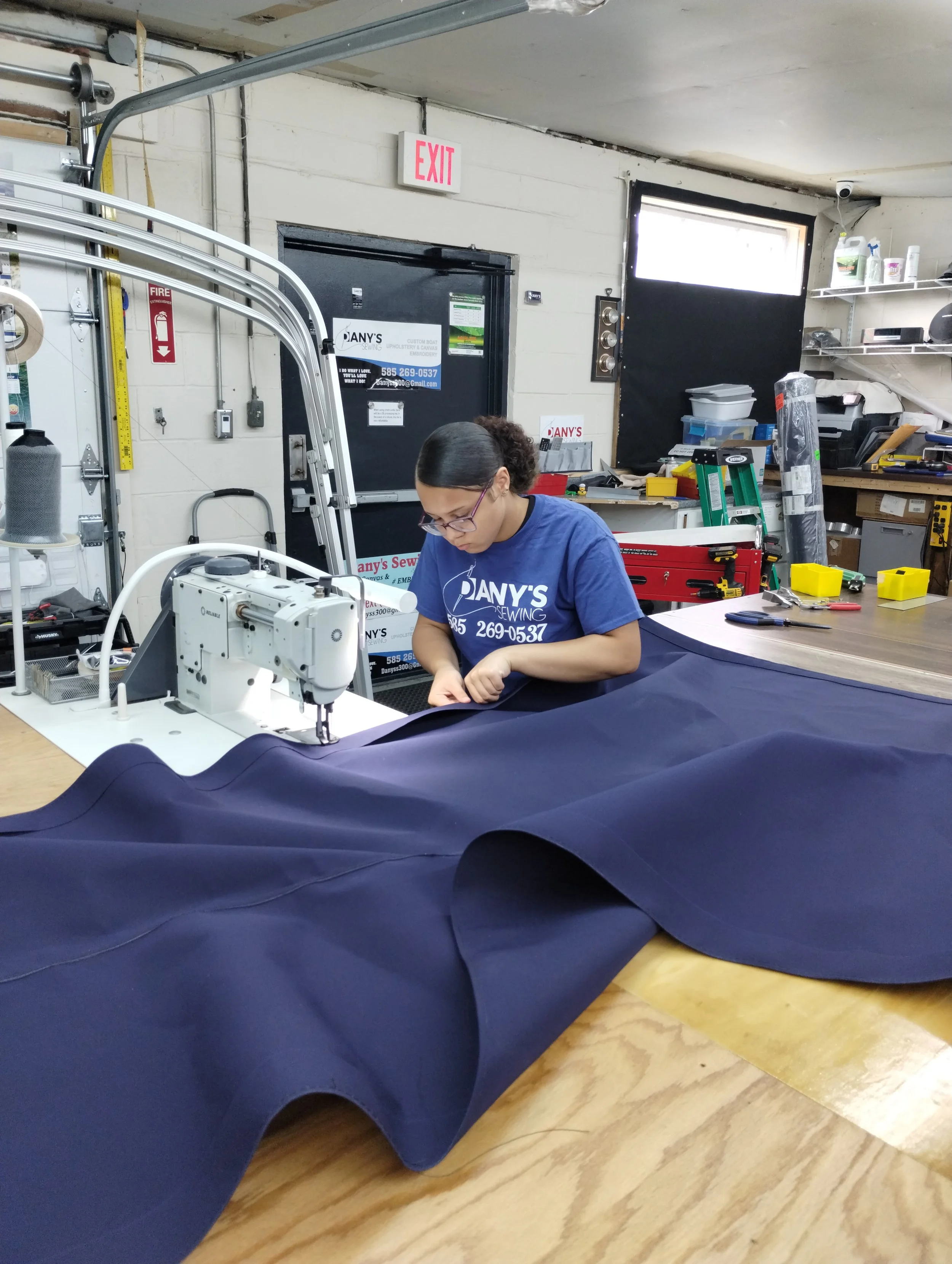 A woman working on a sewing machine in a workshop, surrounded by various tools and supplies, with a dark blue fabric spread out in front of her.