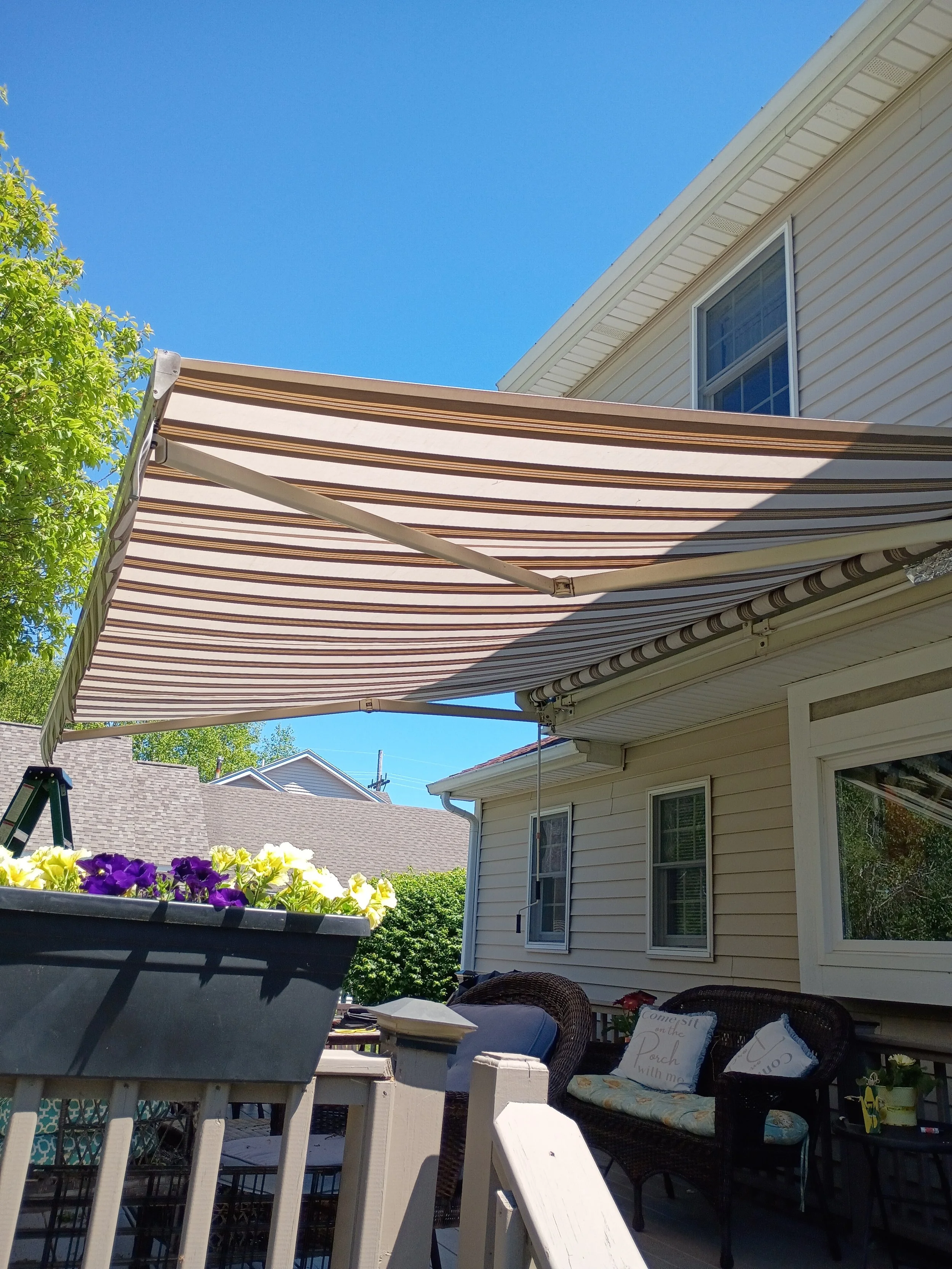 Outdoor patio with striped retractable awning, white house with windows, potted flowers, patio furniture with cushions, and a green tree under a clear blue sky.