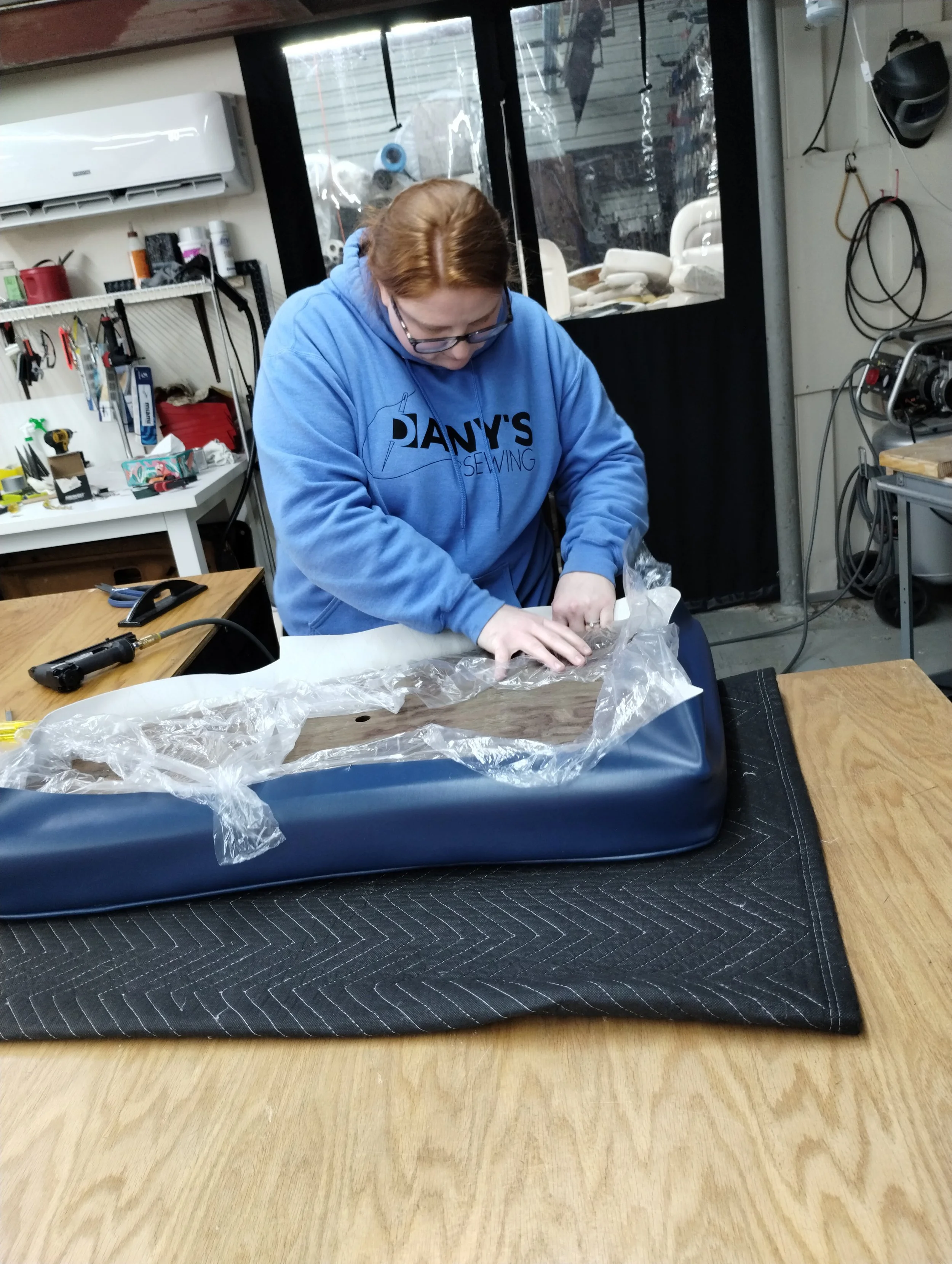 A person with red hair wearing glasses and a blue hoodie working on a craft project in a workshop. The workspace has tools on the shelves and a window with an outdoor view.