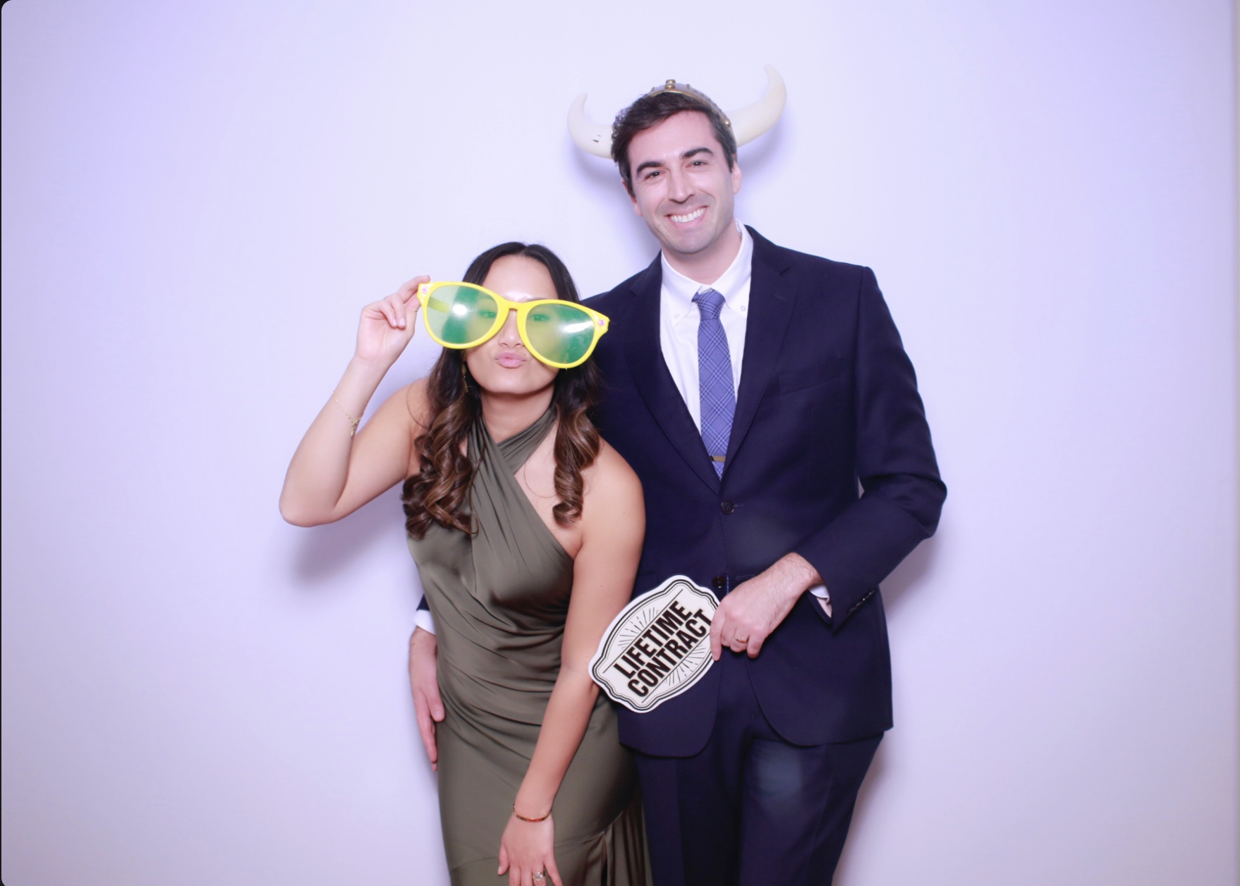 A man and woman stand together against a white background, smiling. The woman is wearing large yellow sunglasses and a green dress, while the man is in a dark suit with a blue tie. The woman is holding a prop that says 'LIFETIME CONTRACT,' and the ma