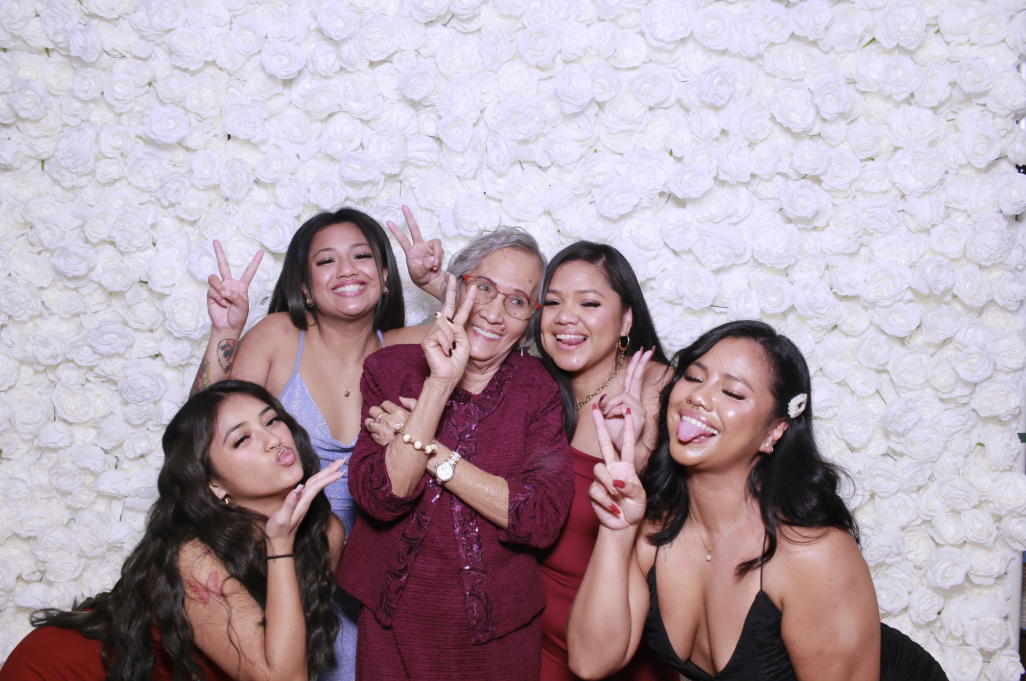 Group of women and an elderly woman posing playfully in front of a wall of white roses.