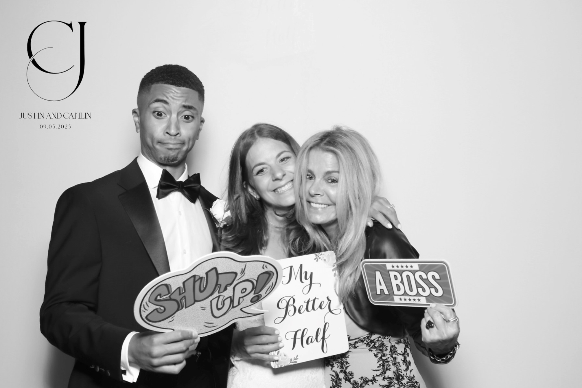 Three people smiling and holding signs. The man on the left is wearing a tuxedo with a bow tie, making a humorous face. The woman in the middle is smiling, holding a sign saying "Shut Up!" The woman on the right has blonde hair, is smiling, and holding a sign that says "A BOSS".