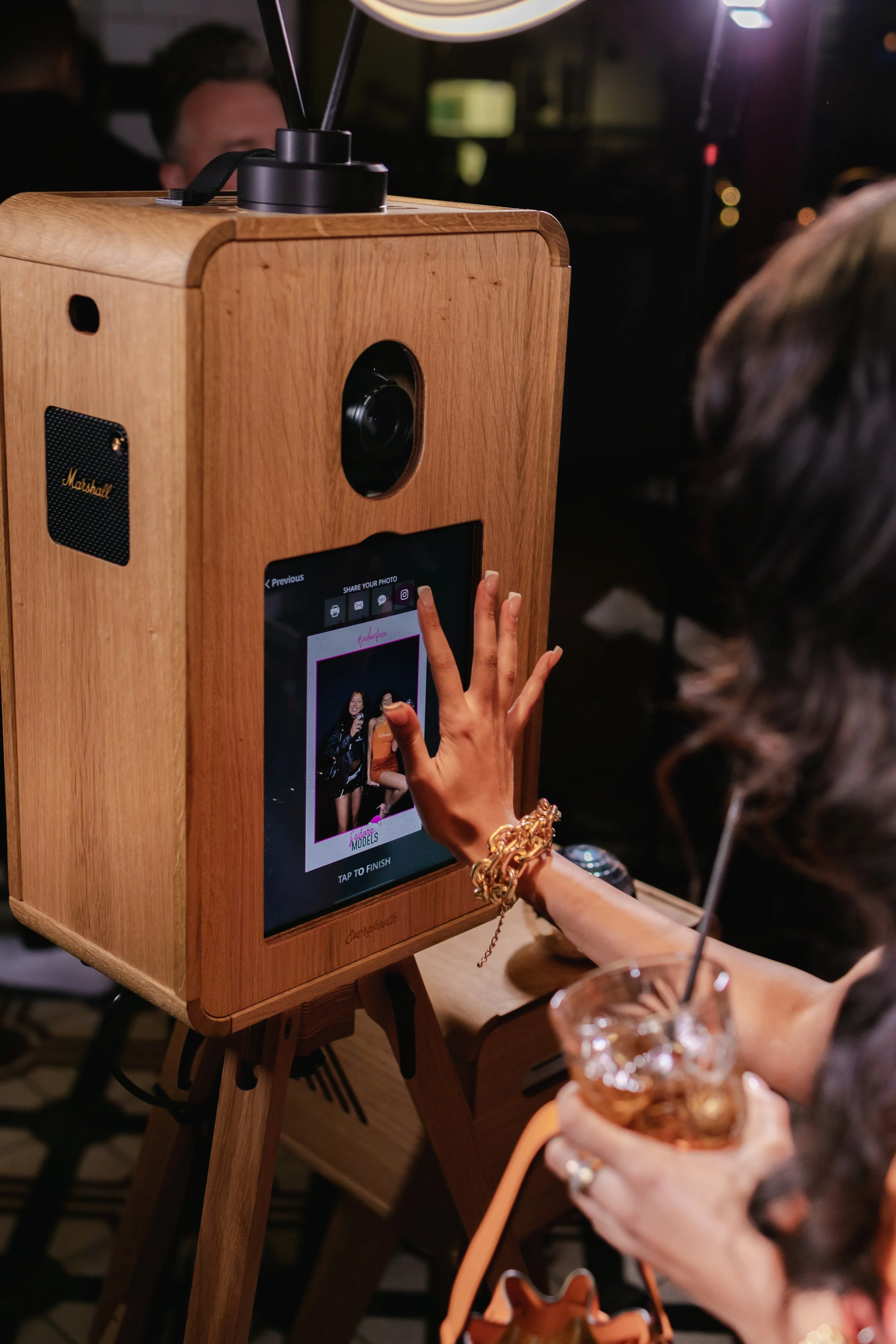 A woman interacts with a photo booth that has a wooden exterior and a touchscreen, capturing a group photo of women. She holds a glass of an iced beverage with a straw and wears jewelry, including bracelets and rings. The photo booth is located in a dark, social setting.