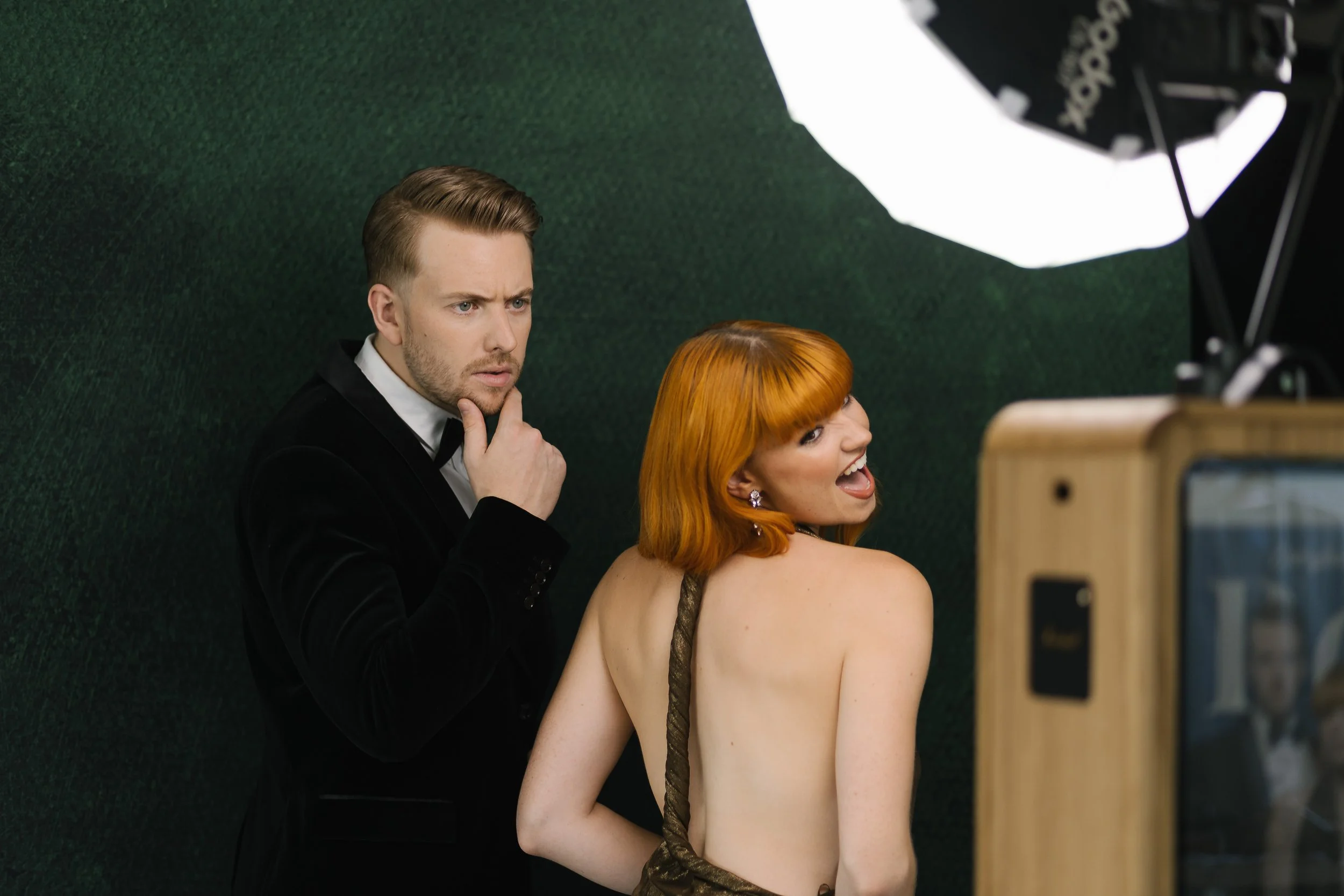 A man in a black tuxedo with a bowtie looks thoughtfully at a woman with bright orange hair who is wearing a gold dress. They are in a photography studio with green textured background and a large softbox light.