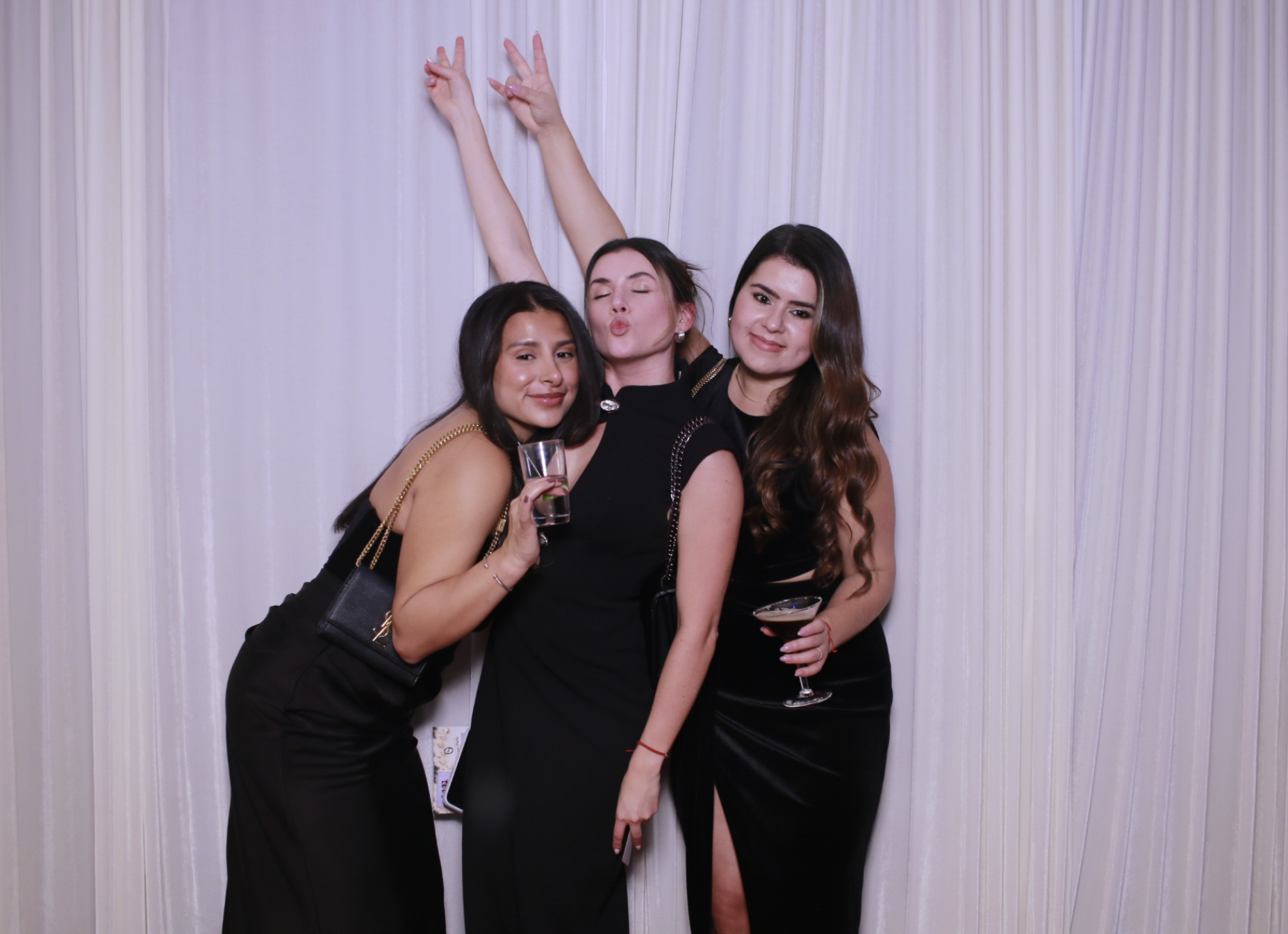Three women in black dresses posing with drinks in front of white curtains, one woman making a kissy face, all smiling and enjoying themselves at what appears to be a party or celebration.