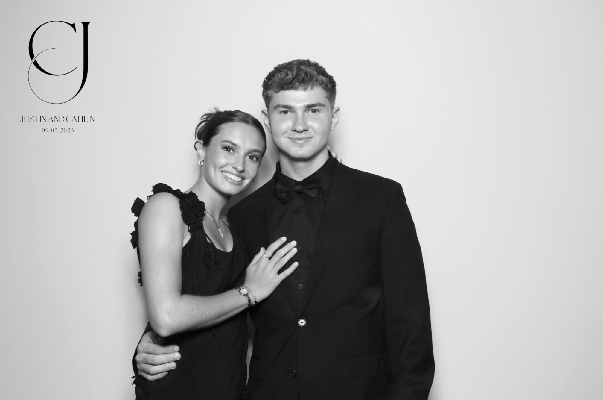 Black and white photo of a smiling couple dressed formally. The woman has her arm around the man, and they are standing close together, looking at the camera. The woman has her hand on the man's chest. The image includes a logo with intertwined lette