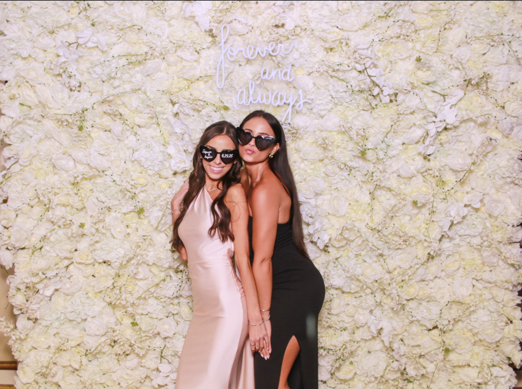 Two women pose in front of a white floral backdrop with a sign that says "forever and always". Both women are wearing black sunglasses, one in a champagne-colored satin dress and the other in a black dress.