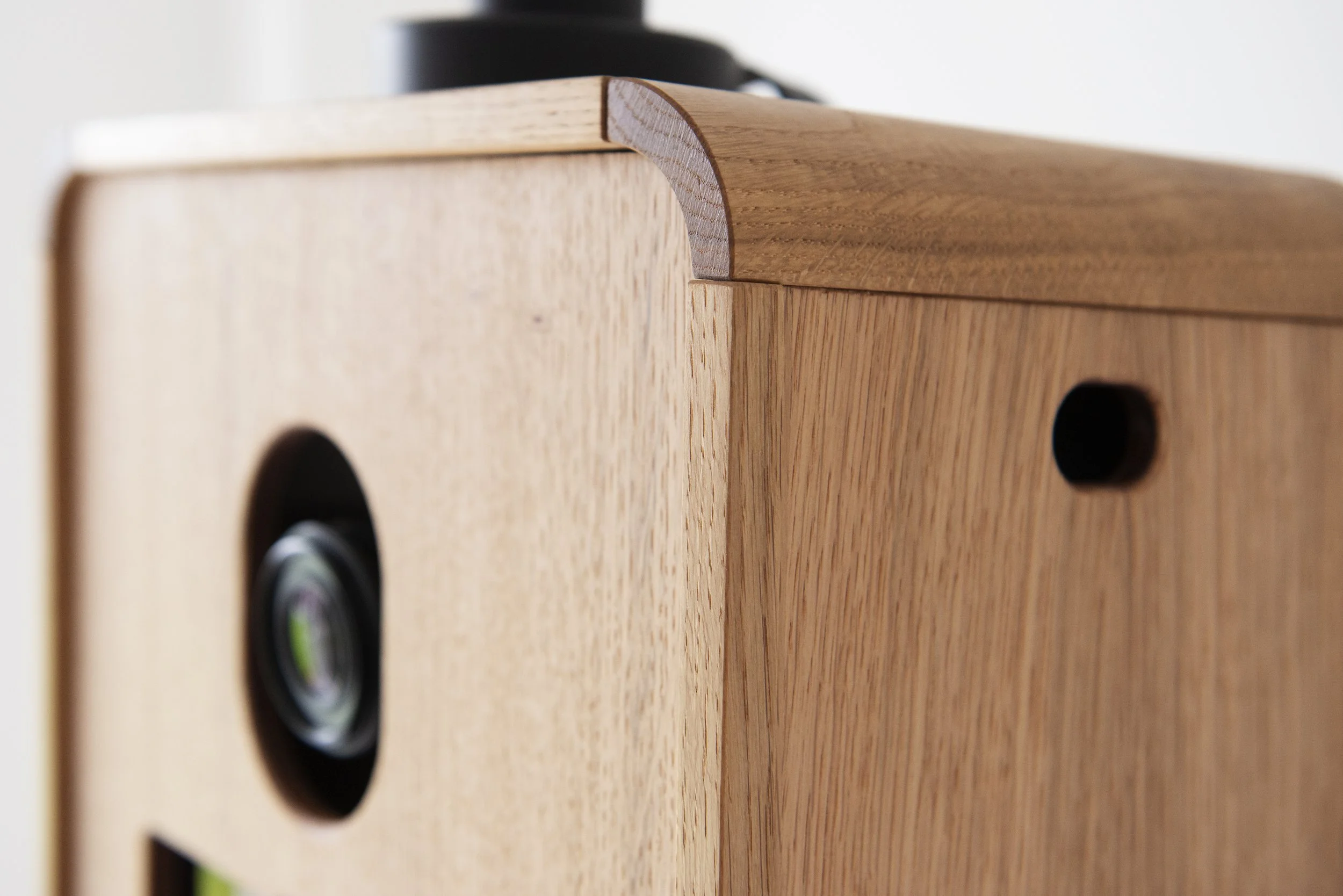 Close-up view of a wooden speaker with a black circular speaker cone and a small black port on the side, showing detailed wood grain and smooth edges.