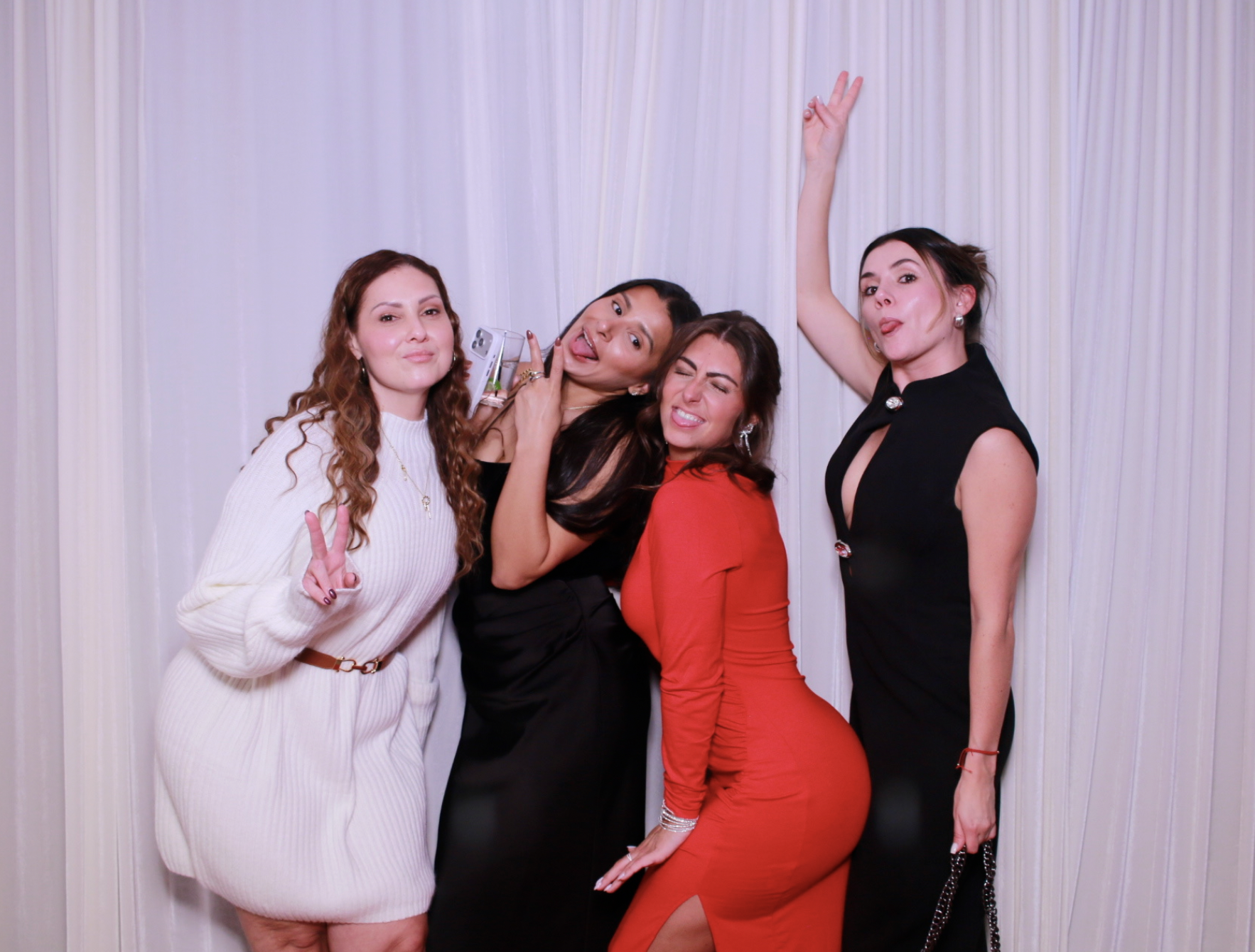 Four women posing playfully in front of a white curtain. They are making various facial expressions and hand gestures, with some sticking out tongues, smiling, and peace signs.