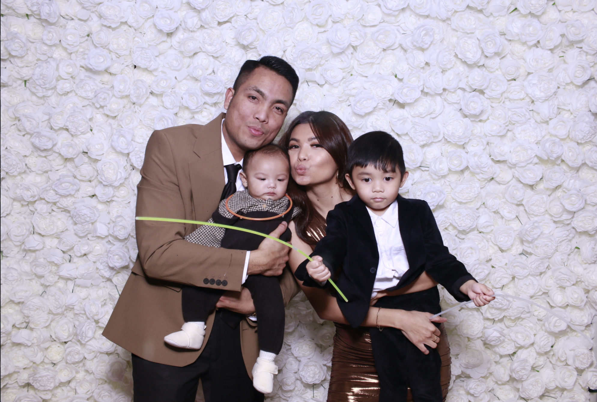 A family of four posing in front of a white flower wall, with a man in a brown suit, woman in a shiny copper dress, and two children, one boy in a black blazer and white shirt, and a girl in a dress holding a green balloon.
