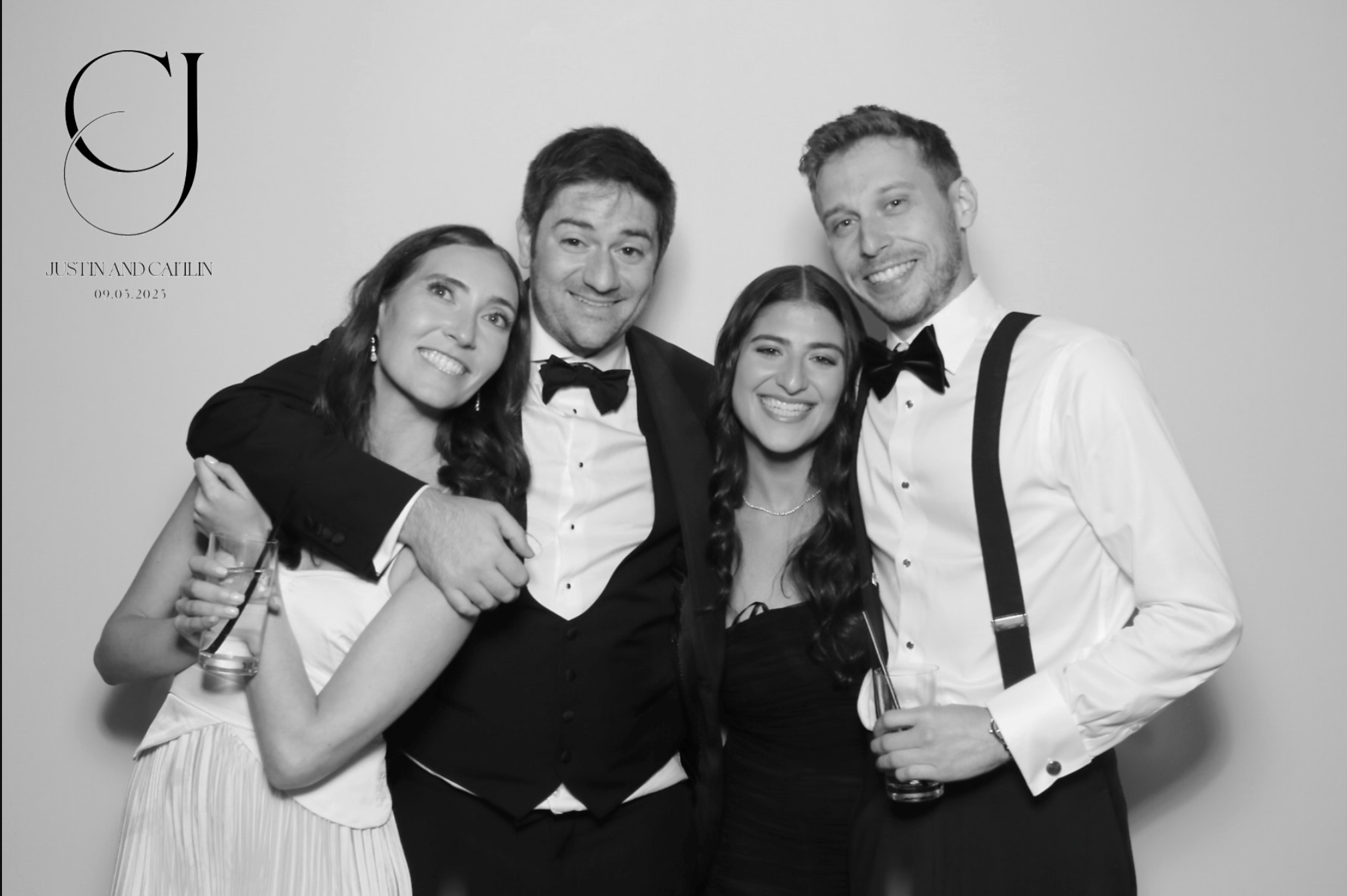 Black and white photo of four people dressed in formal attire, smiling, with two women and two men. The woman on the left is holding a drink and has dark hair, the man in the middle has dark hair, the woman next to him has dark wavy hair, and the man